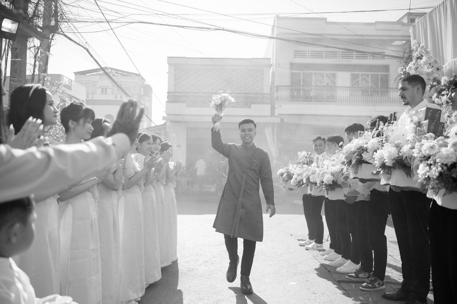 A figure in a coat walks between rows of spectators with raised hands in a black and white scene.