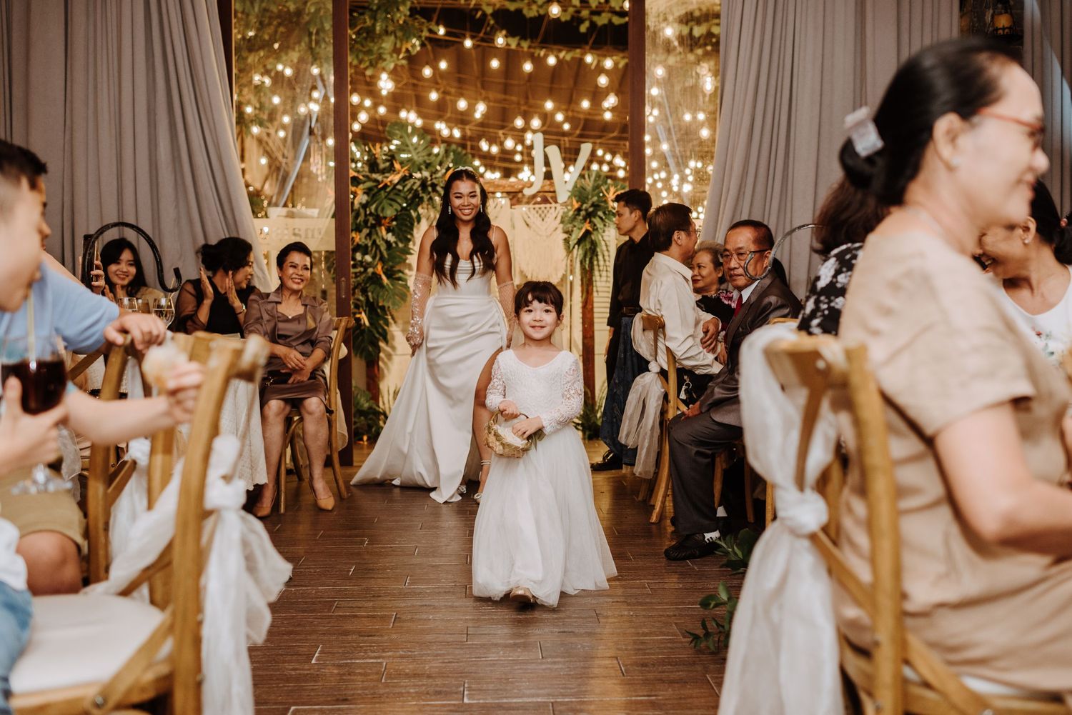 Young ring bearer walking down aisle at indoor wedding ceremony decorated with string lights and floral arch.