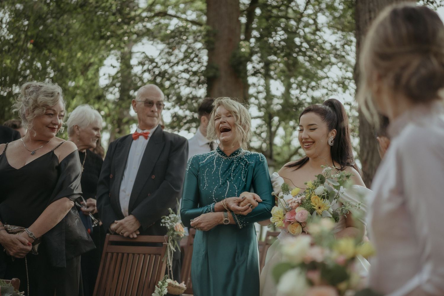 Wedding guests share joyful laughter during an outdoor ceremony among trees.