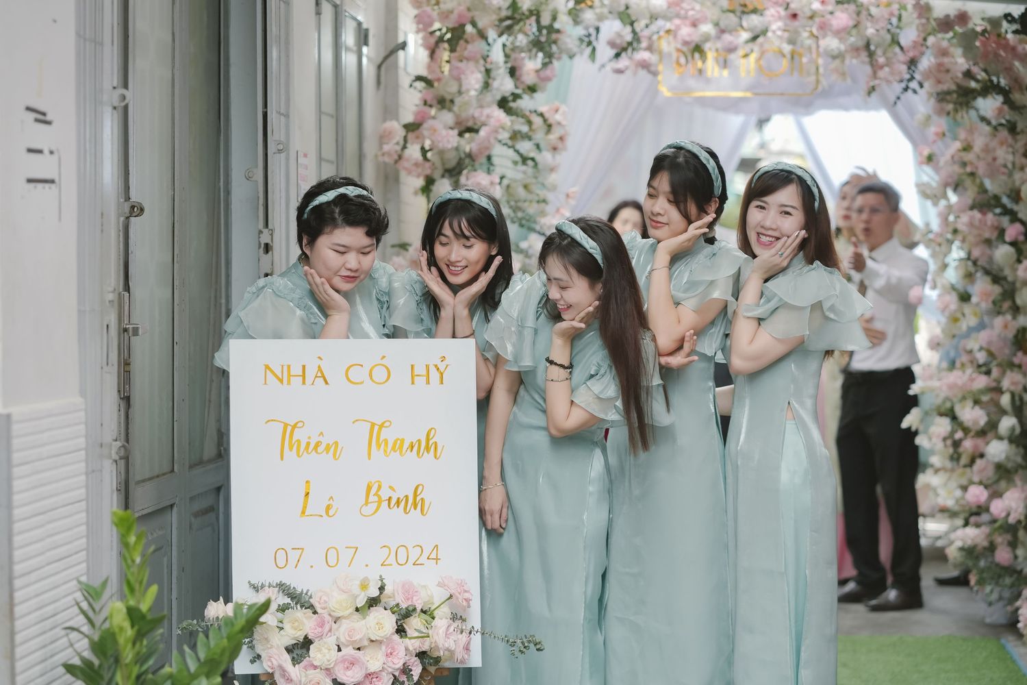 Wedding party in light blue dresses poses with welcome sign under floral arch.