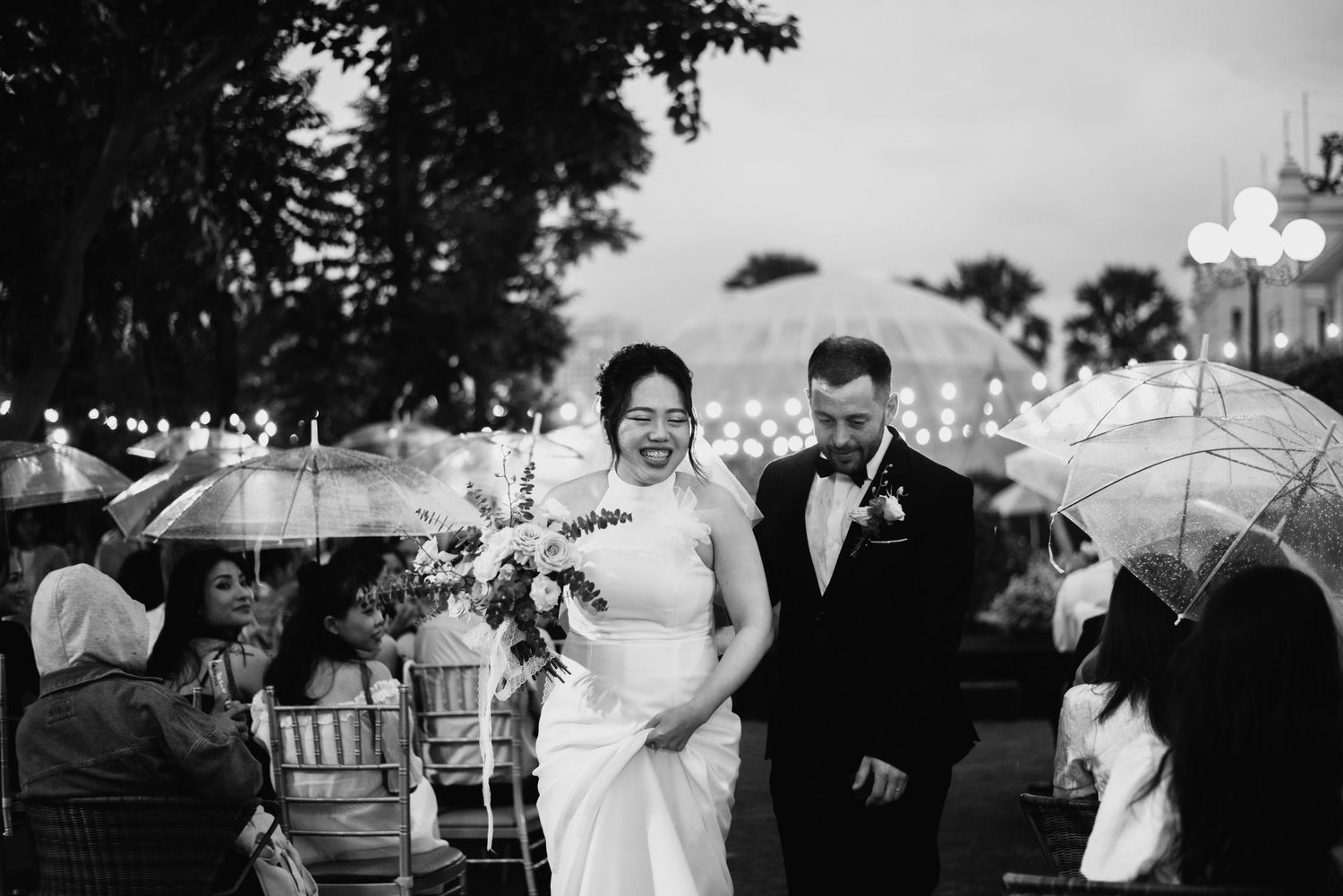 A rainy wedding celebration with guests holding clear umbrellas during an outdoor ceremony.