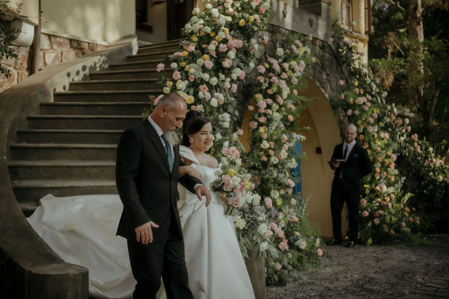 A wedding couple descends elegant stone stairs adorned with lush white floral arrangements.