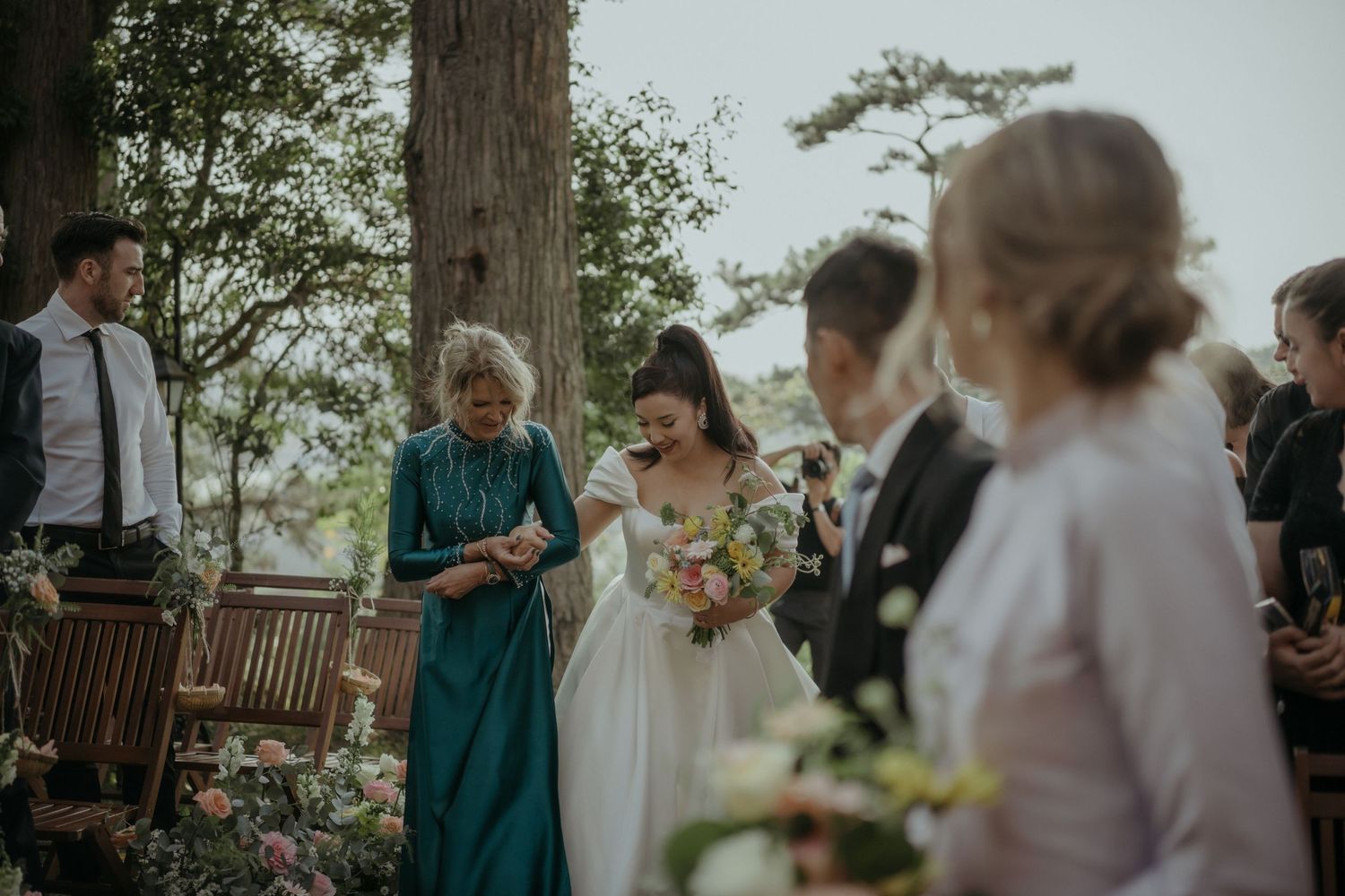 An intimate outdoor wedding ceremony takes place near trees with attendees gathered around a wooden bench.