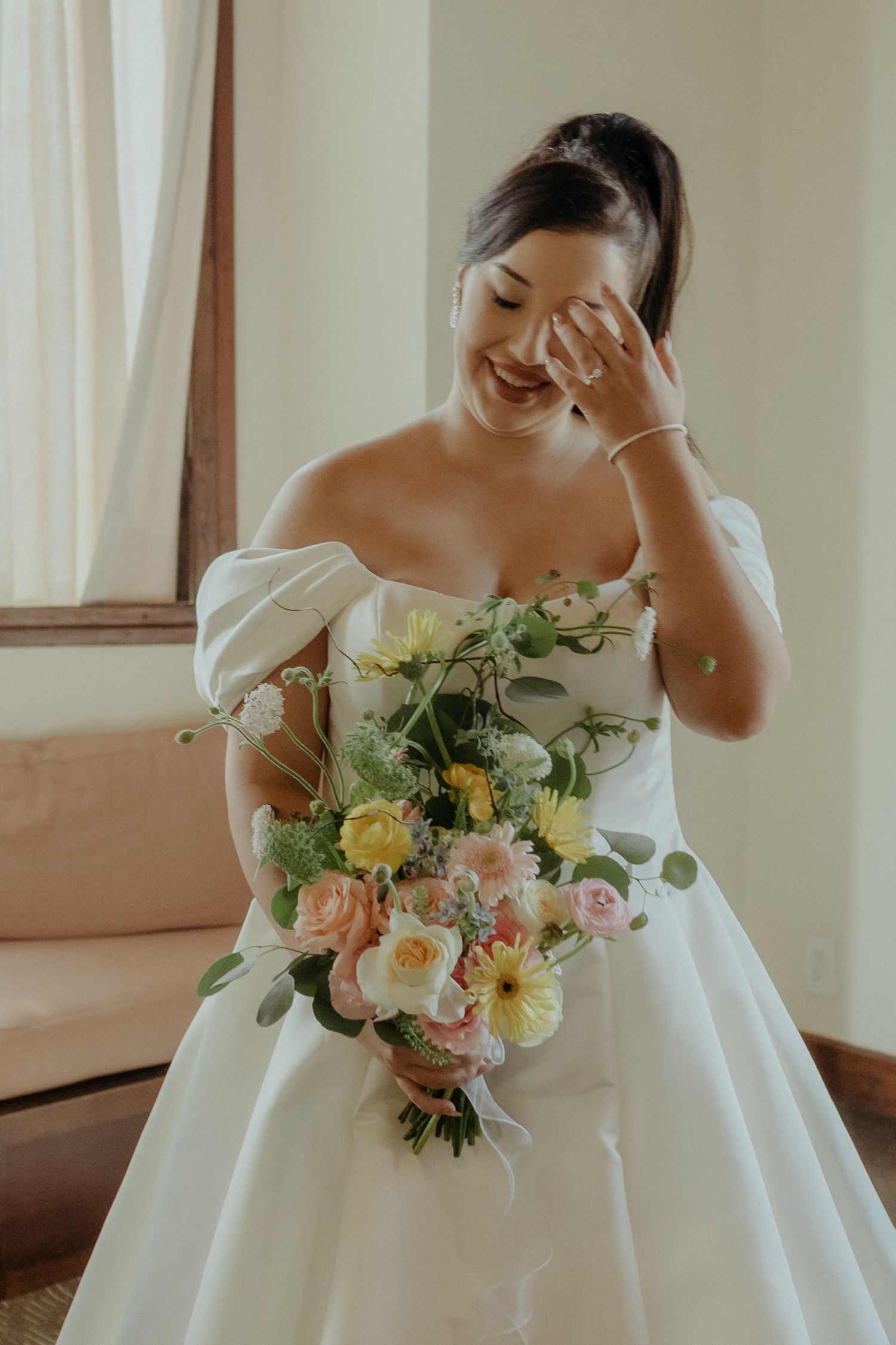 A bride in an elegant white off-shoulder wedding gown laughs joyfully while holding a pastel bouquet of roses and greenery.