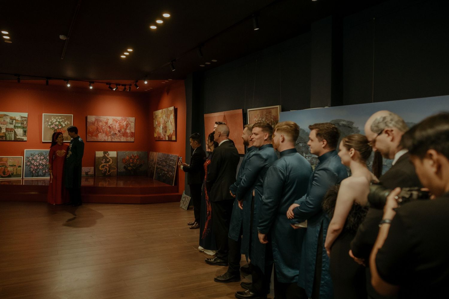 A line of people standing in an art gallery with red walls and dimly lit lighting viewing framed artworks on display.