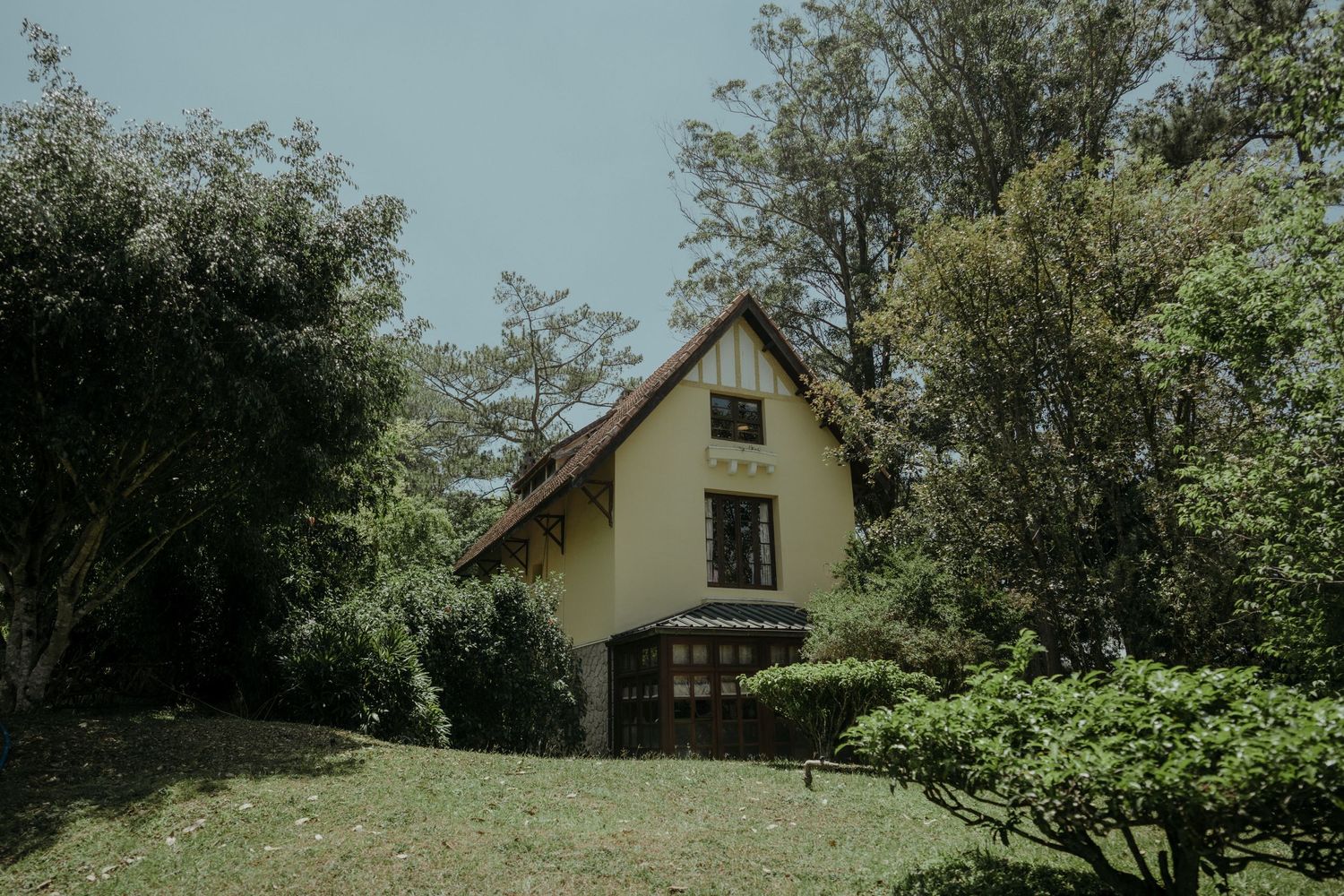 A charming yellow cottage with an A-frame design sits nestled among lush green trees and vegetation on a sunny day.