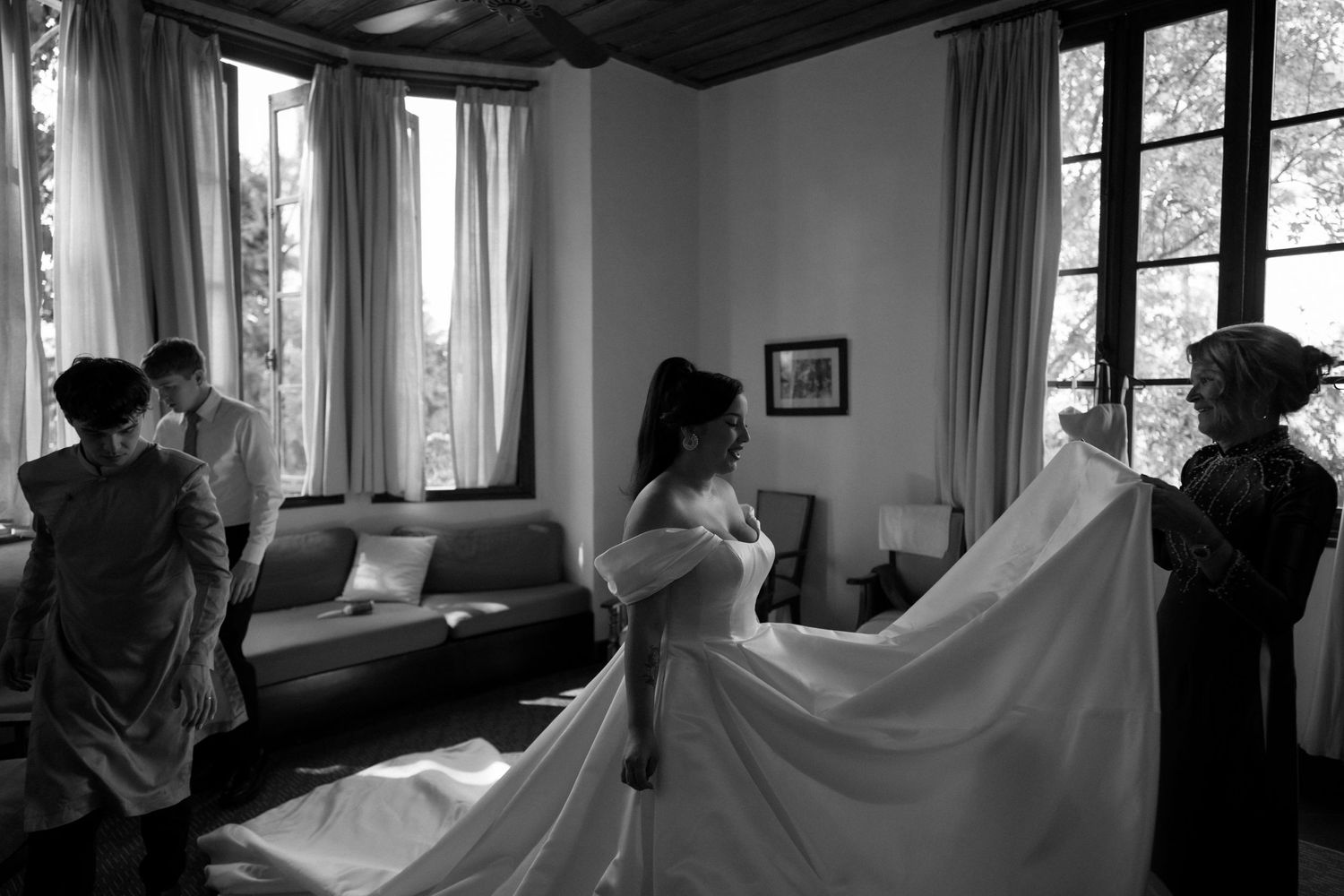 Black and white photo of people helping someone get dressed in a dress with a long flowing train in a sunlit room.