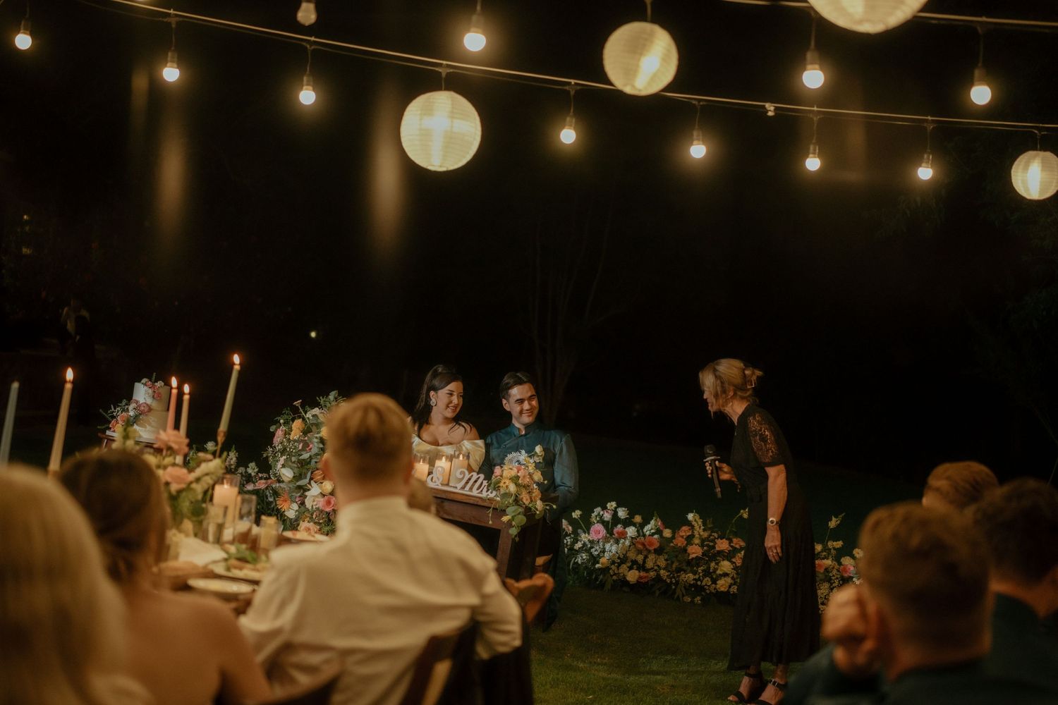 An outdoor evening reception with string lights and paper lanterns illuminating a dinner party under the night sky.