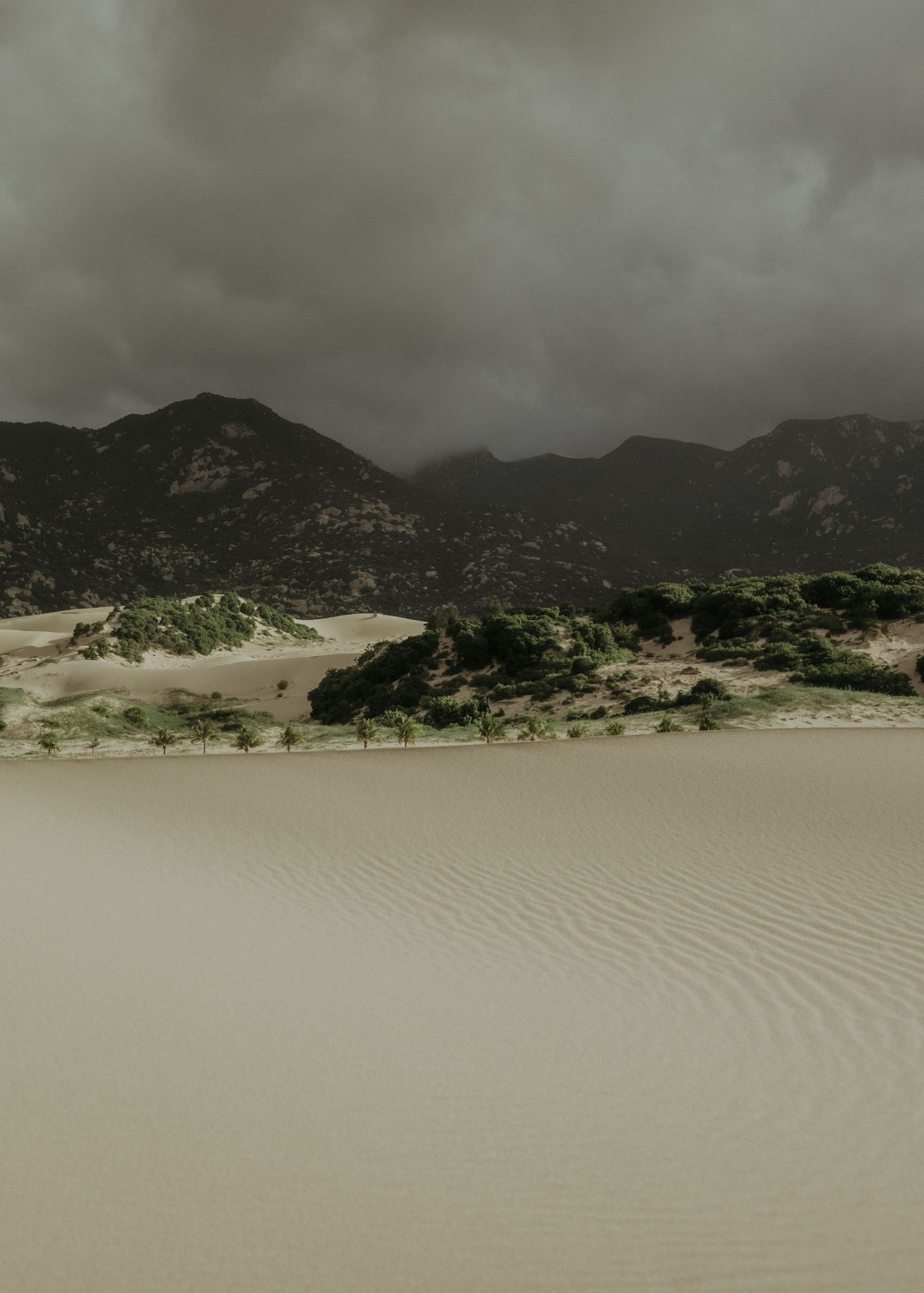 Dramatic storm clouds gather over sand dunes and mountains in a moody desert landscape.