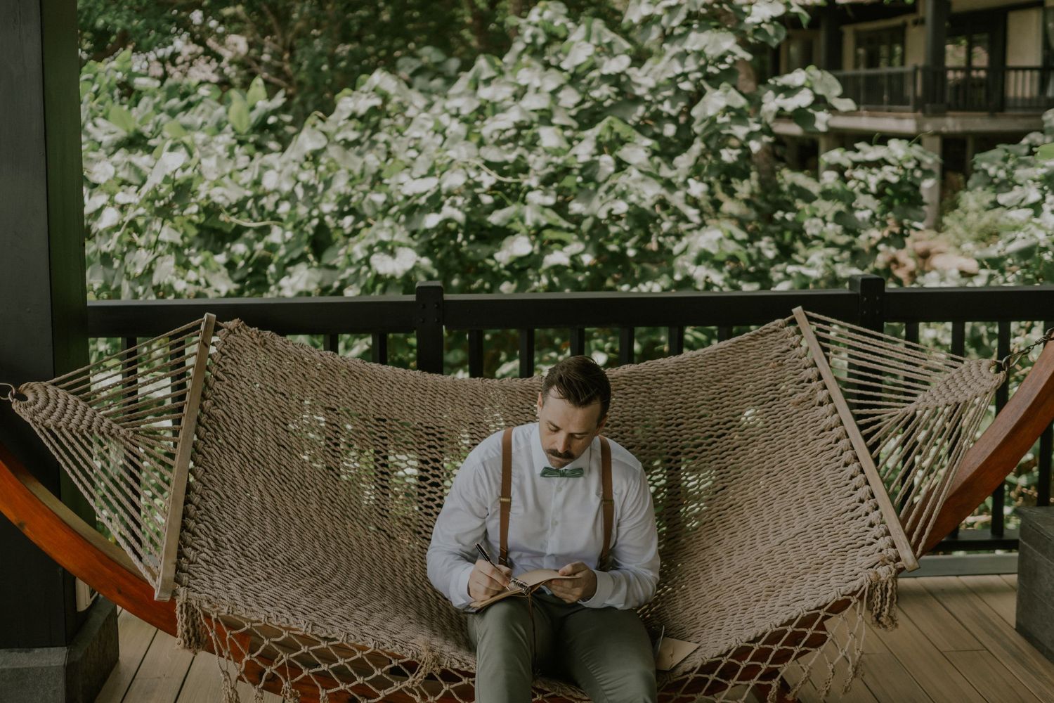 Person relaxing in a woven hammock on a wooden porch surrounded by lush greenery.