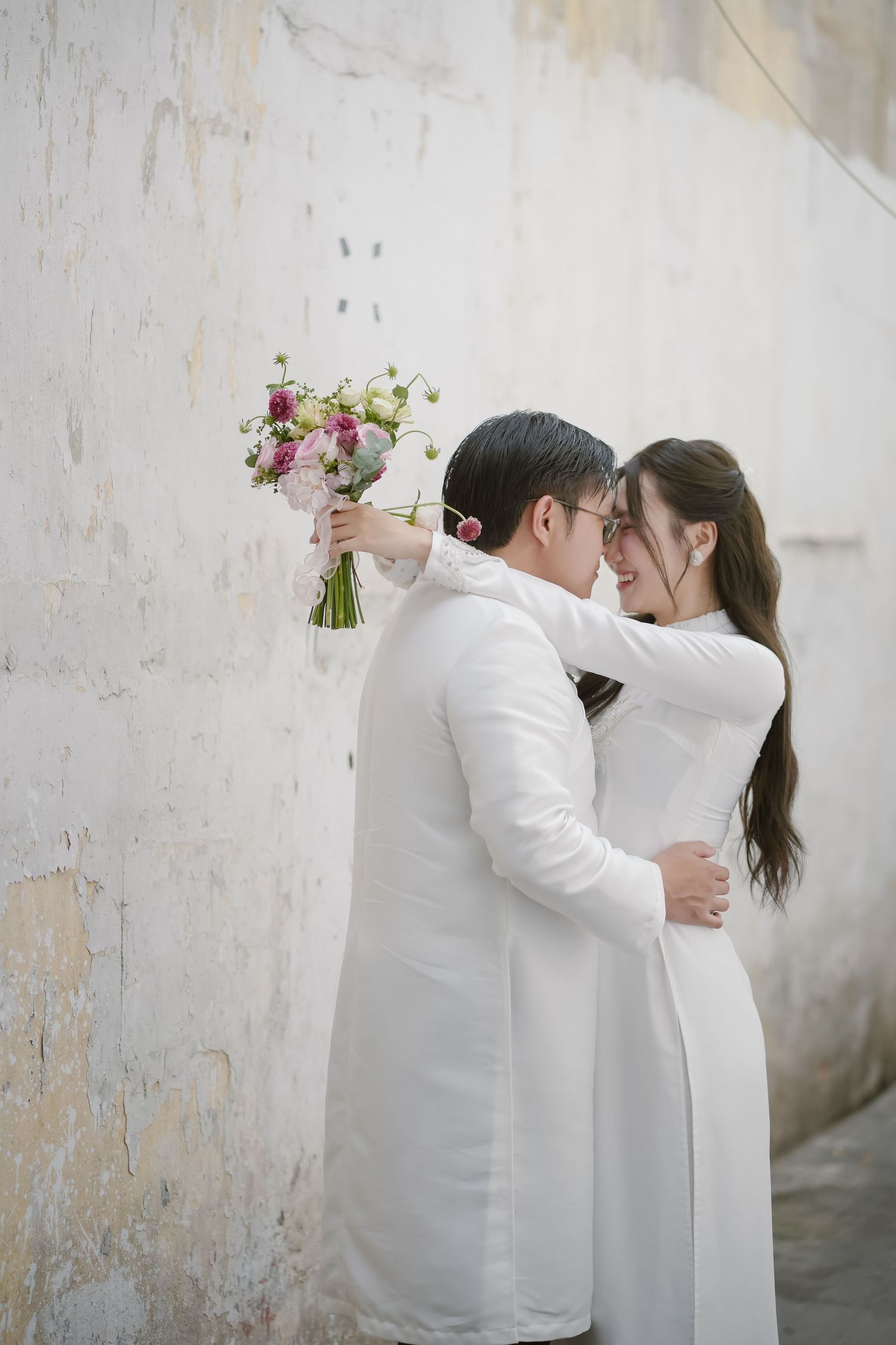 Intimate moment between couple in white attire holding pink bouquet against textured wall.