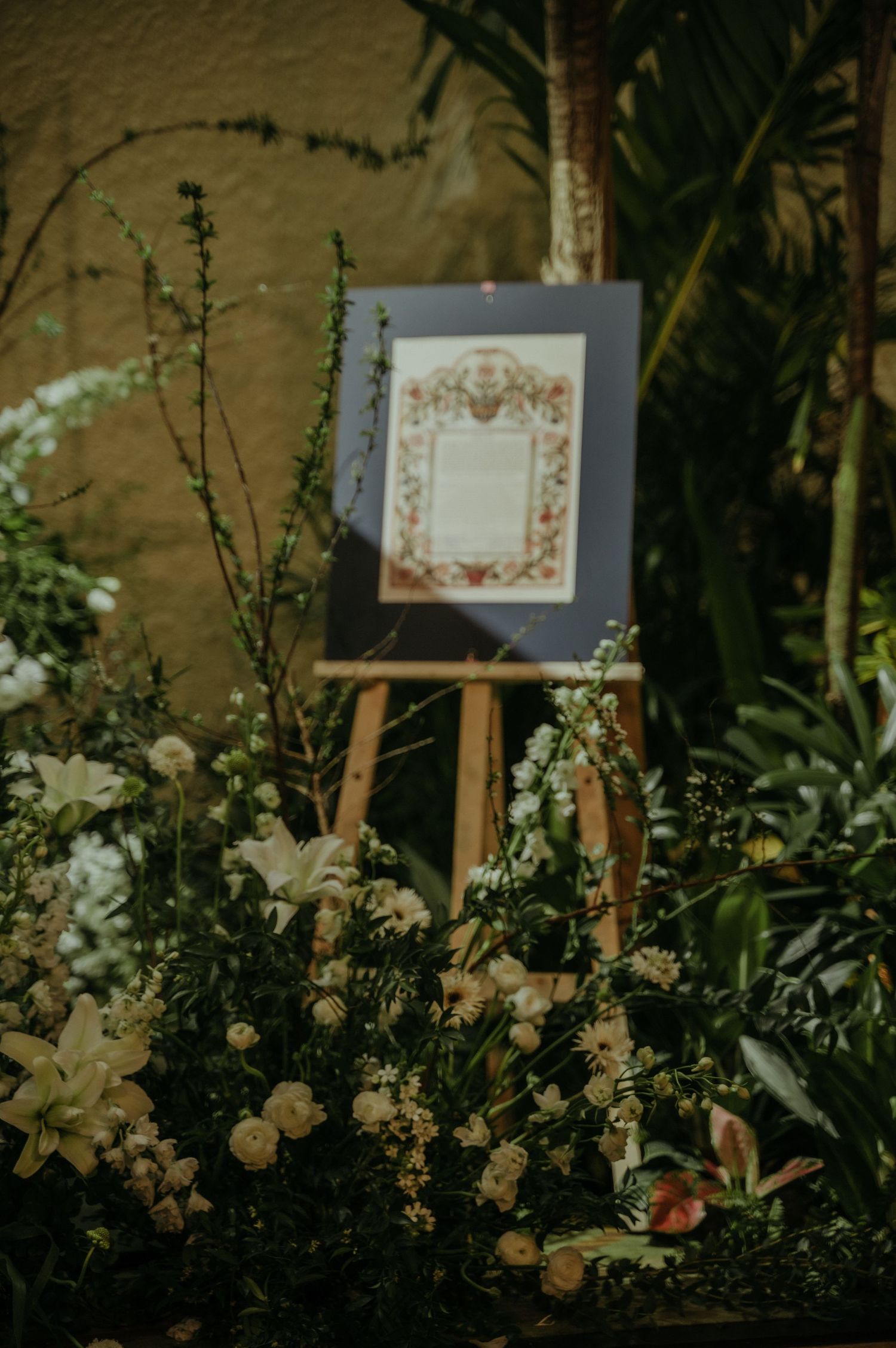 Ornate framed sign displayed on easel amid lush floral arrangements.