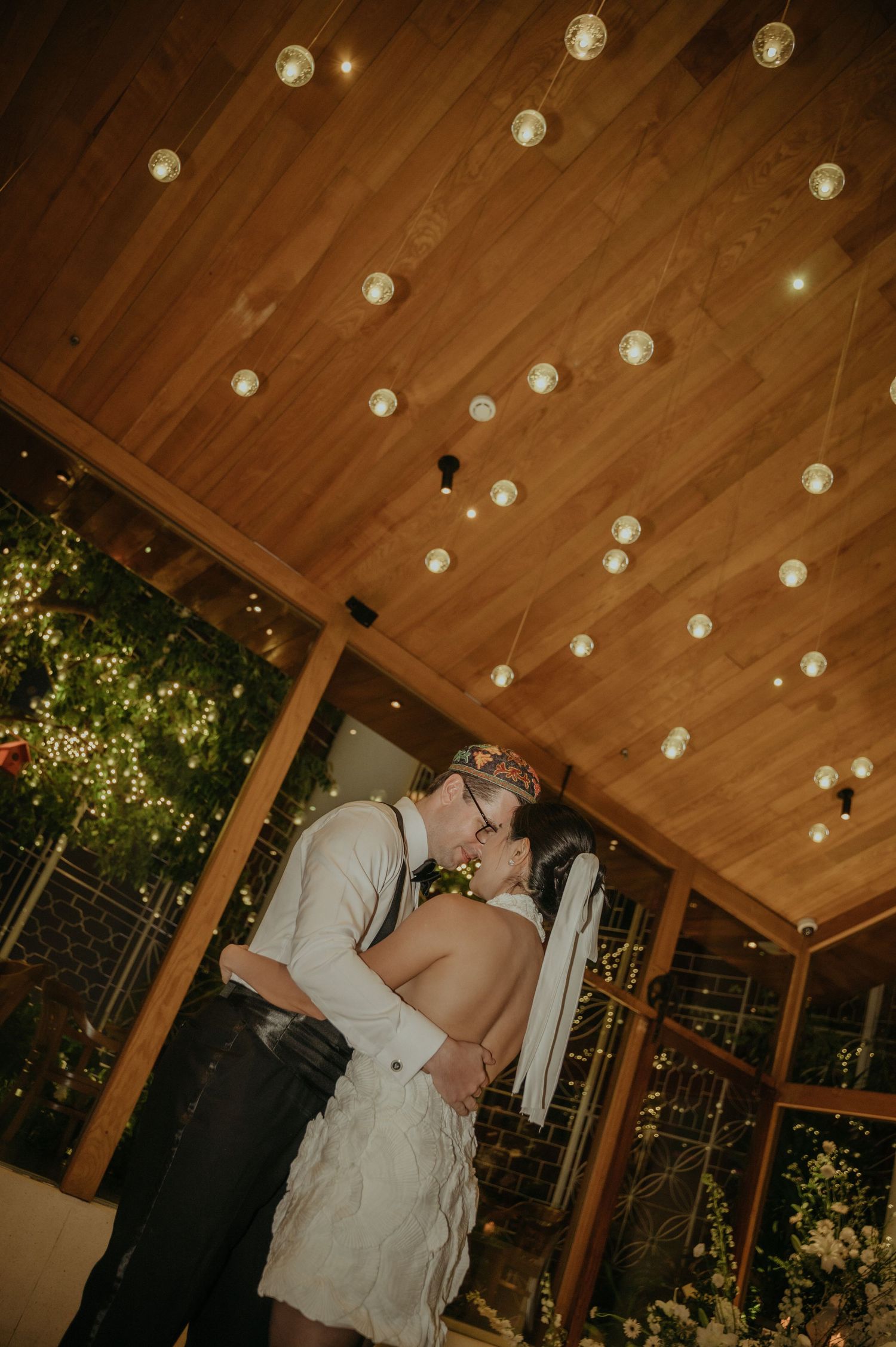 Romantic kiss under wooden ceiling with string lights at wedding reception.