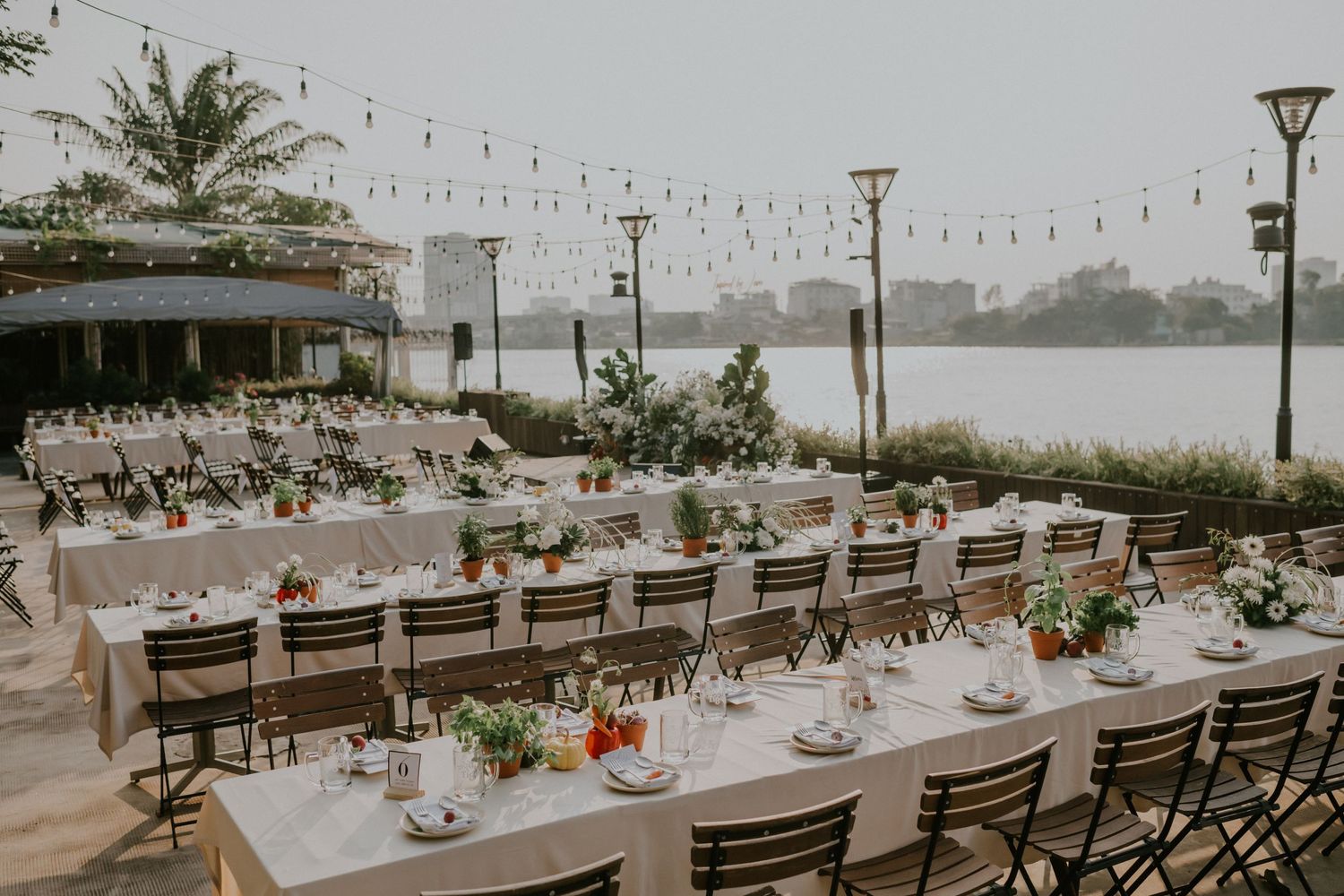 Long dining tables with metal chairs set for outdoor waterfront reception under string lights.
