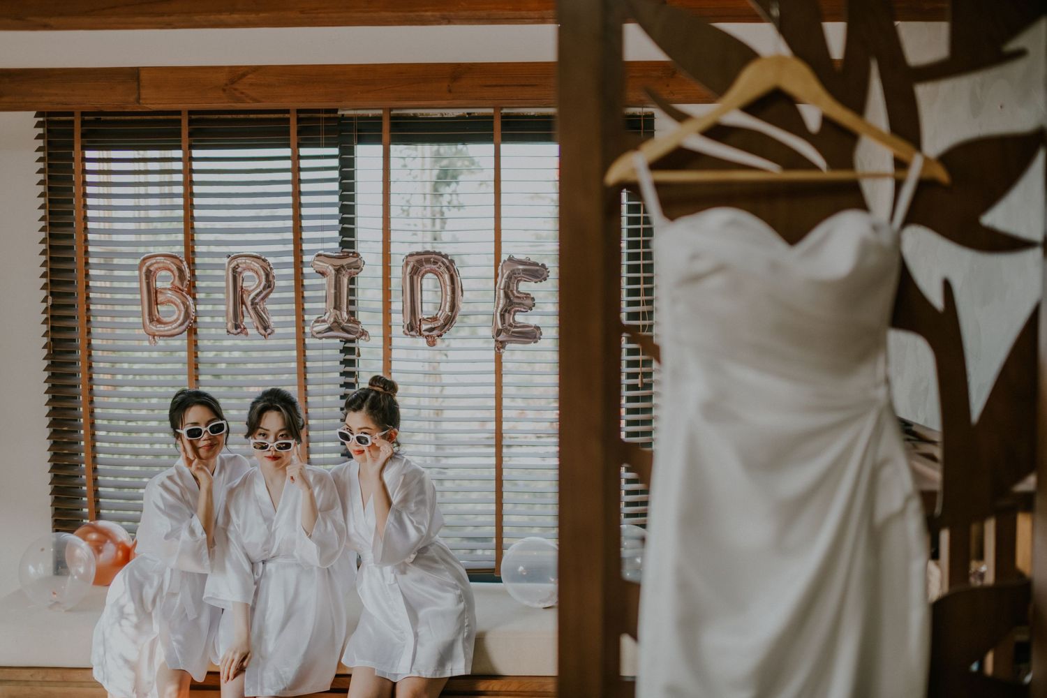 Bridesmaids in white robes pose together with a wedding dress hanging in the background during pre-ceremony preparations.