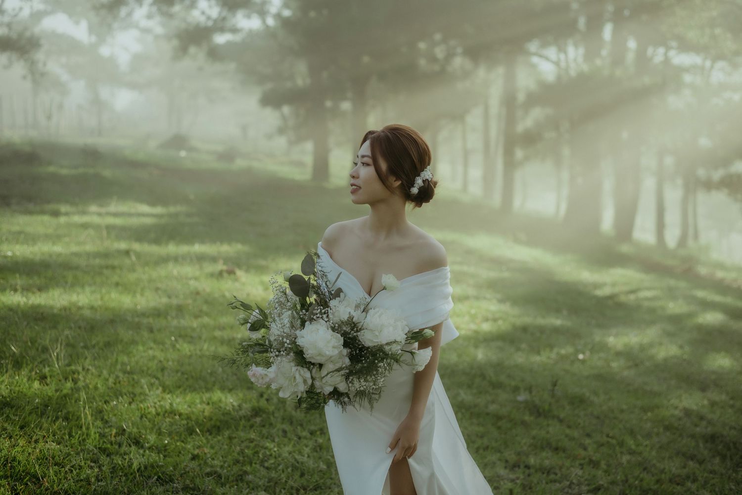 A bride in an off-shoulder white dress holds a lush white bouquet in a misty forest setting during golden hour.