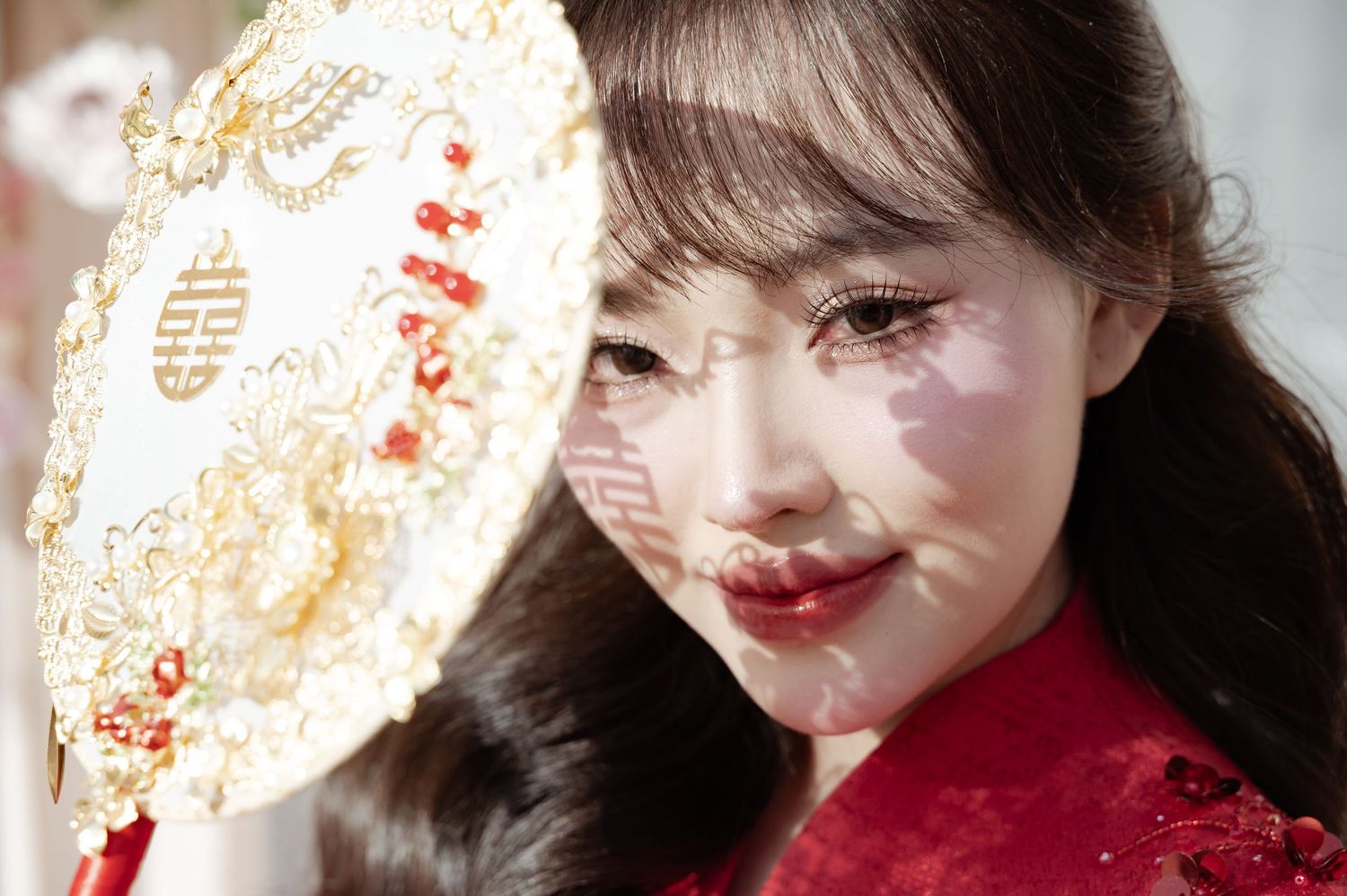 A person in traditional red Chinese attire holds an ornate gold and white paper fan in sunlit setting.
