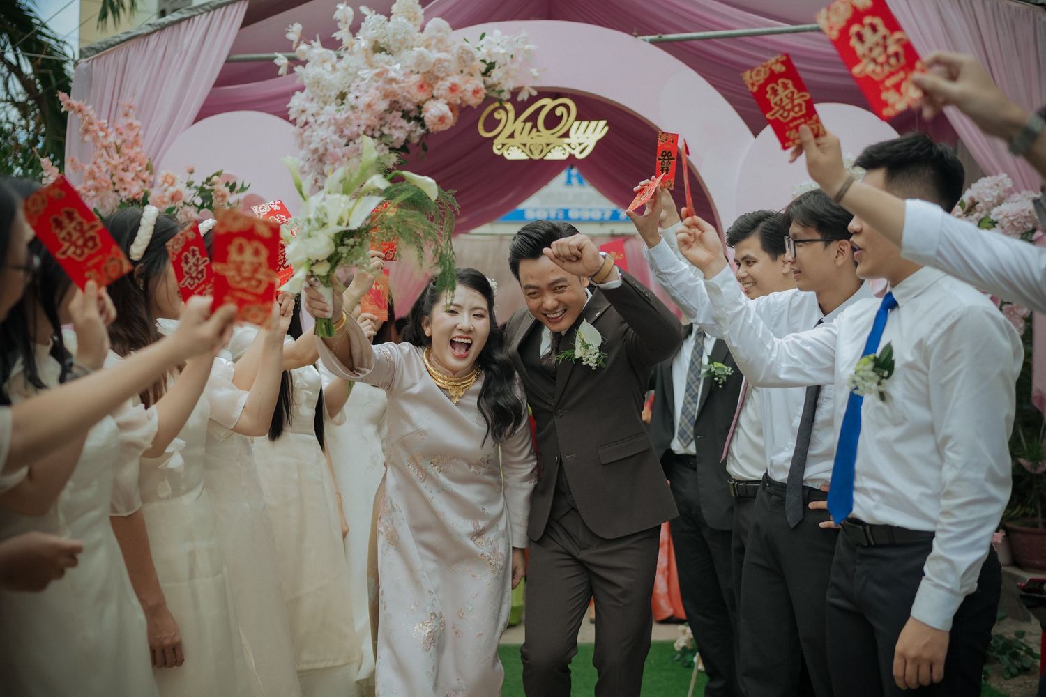 Wedding guests in light colored attire celebrate under an arch decorated with white flowers and red accents.