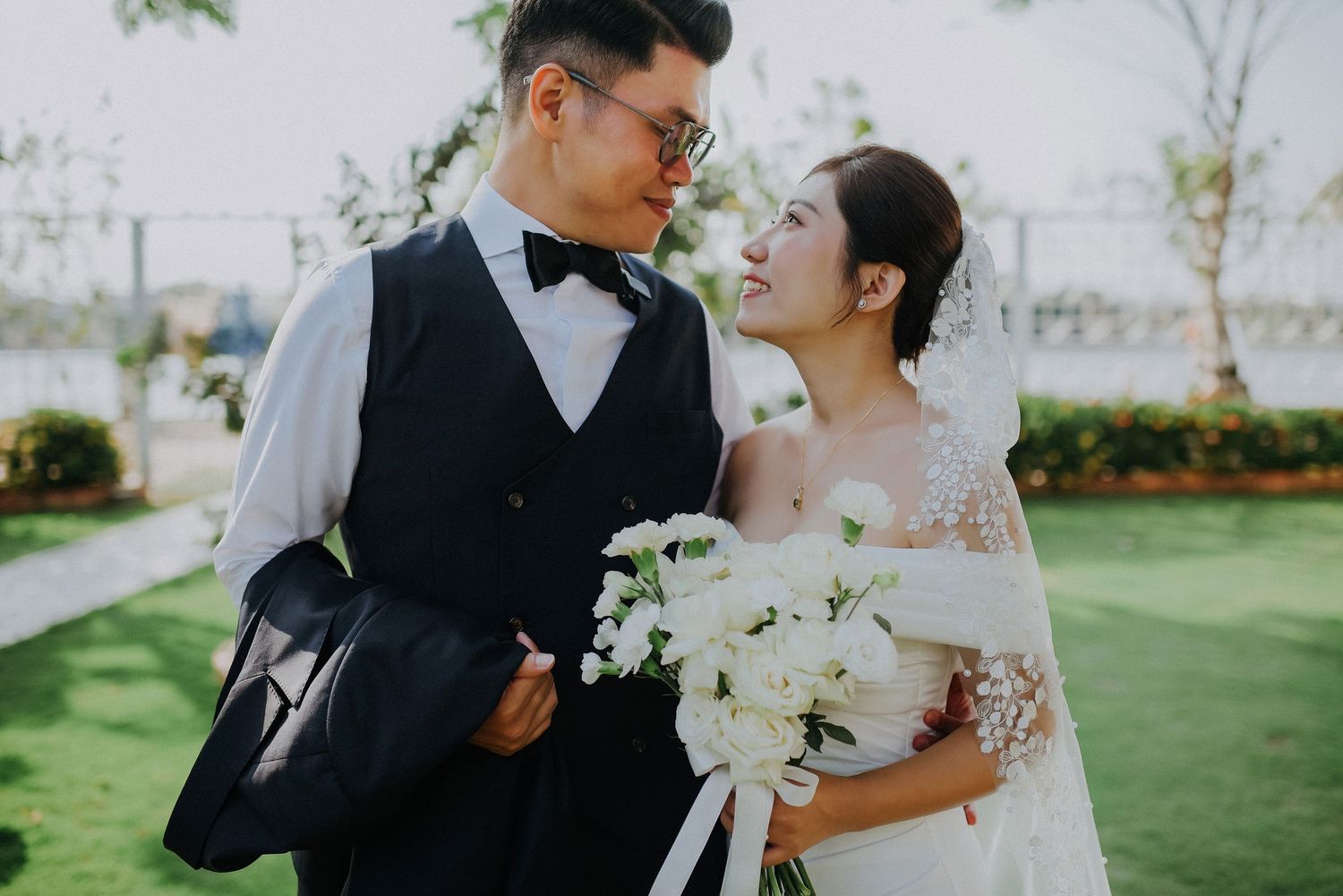 A wedding couple in formal attire poses outdoors on a sunny lawn with white floral bouquet.