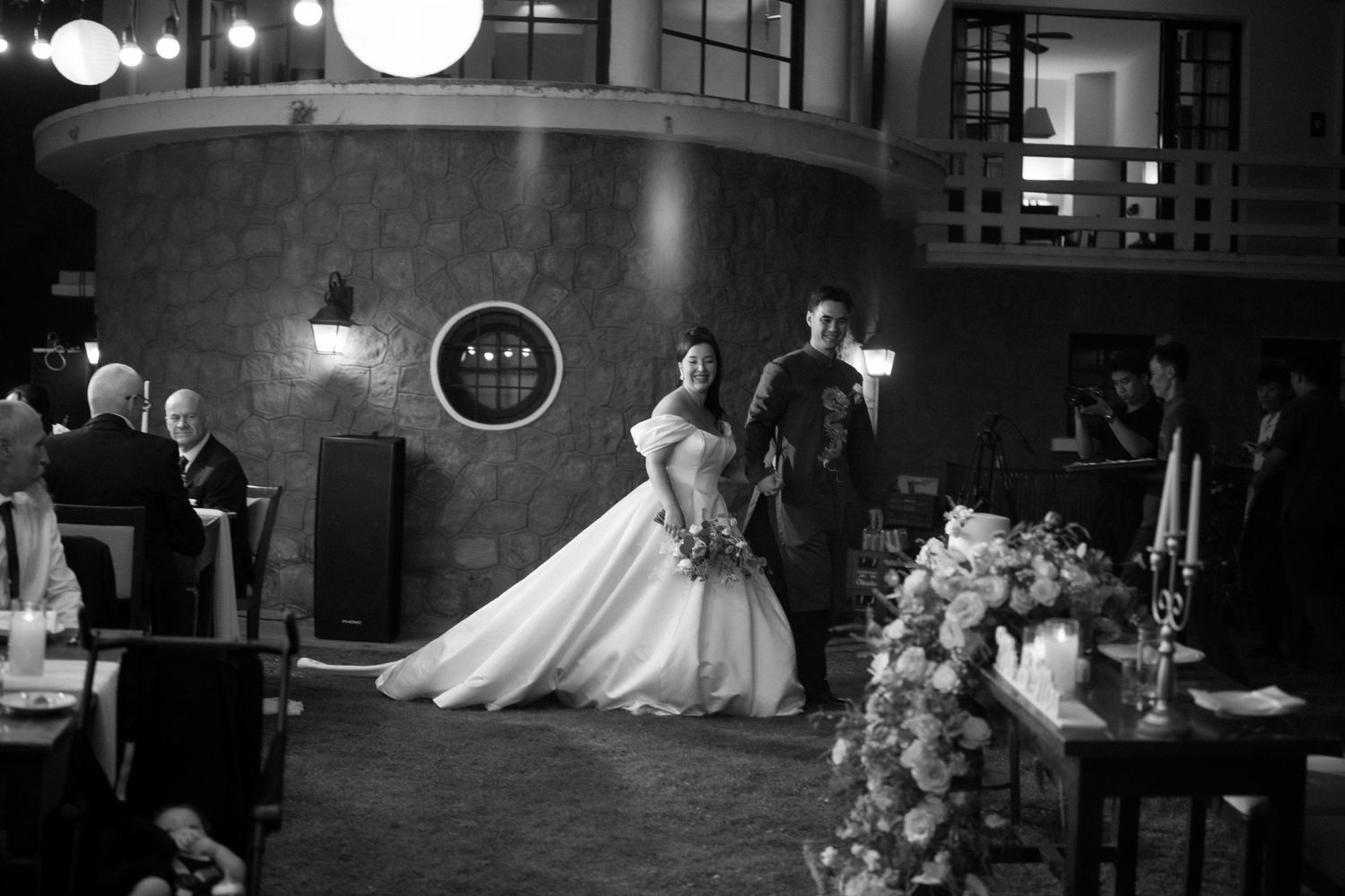 Black and white wedding photograph showing a couple in formal attire dancing in an elegant venue with dramatic lighting.