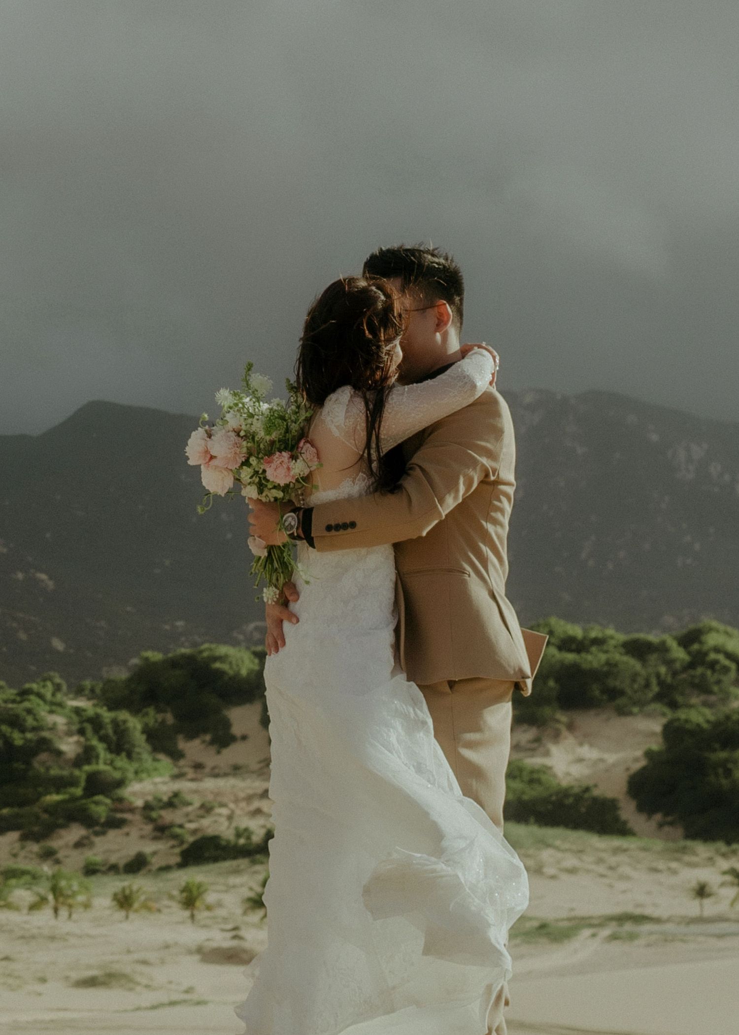 A wedding couple embraces on a beach with mountains and moody clouds in the background.