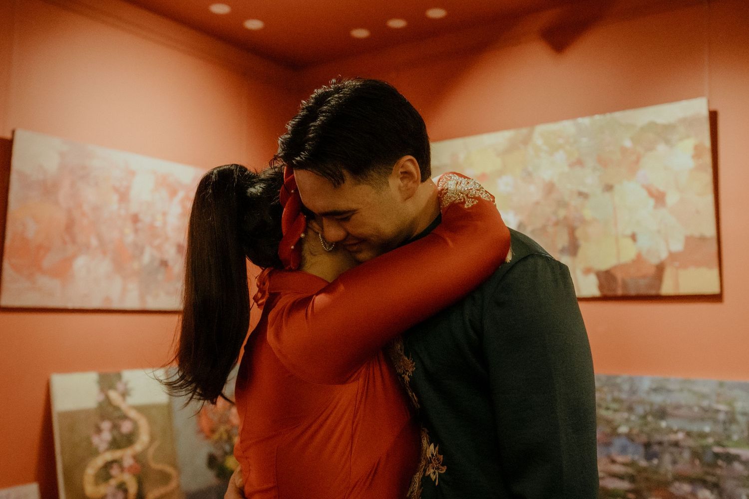 A couple embraces tenderly in a warm-lit room with abstract art on the orange walls behind them.