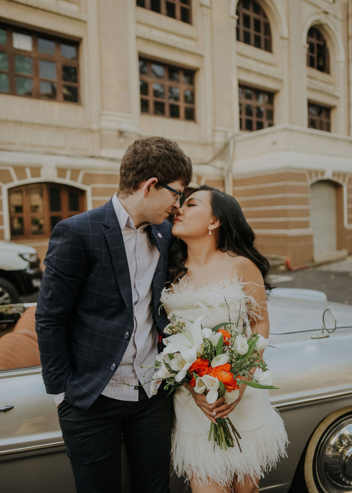 Newlywed couple sharing an intimate moment beside a vintage car outside a historic building.