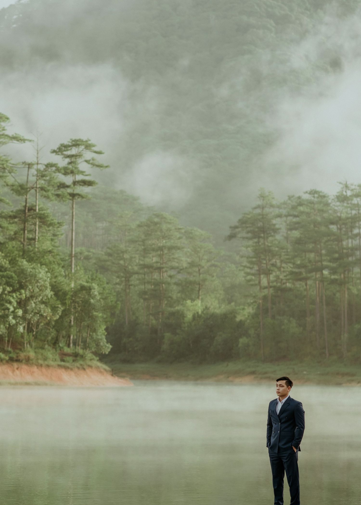 Misty morning by a tranquil lake surrounded by tall pine trees with a figure standing on the shoreline in a dark suit.