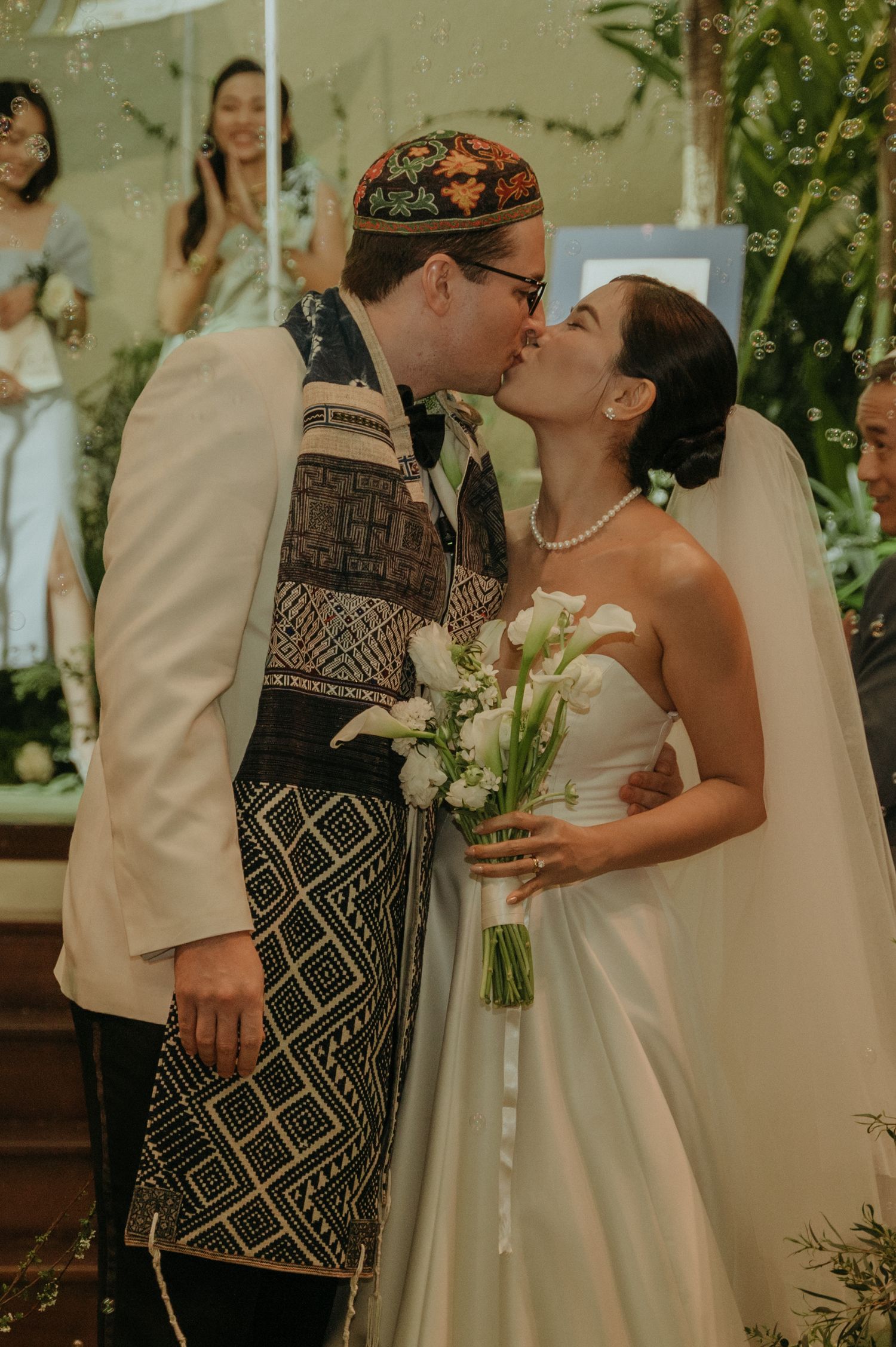 A couple shares a romantic kiss during their wedding ceremony wearing traditional and modern attire.