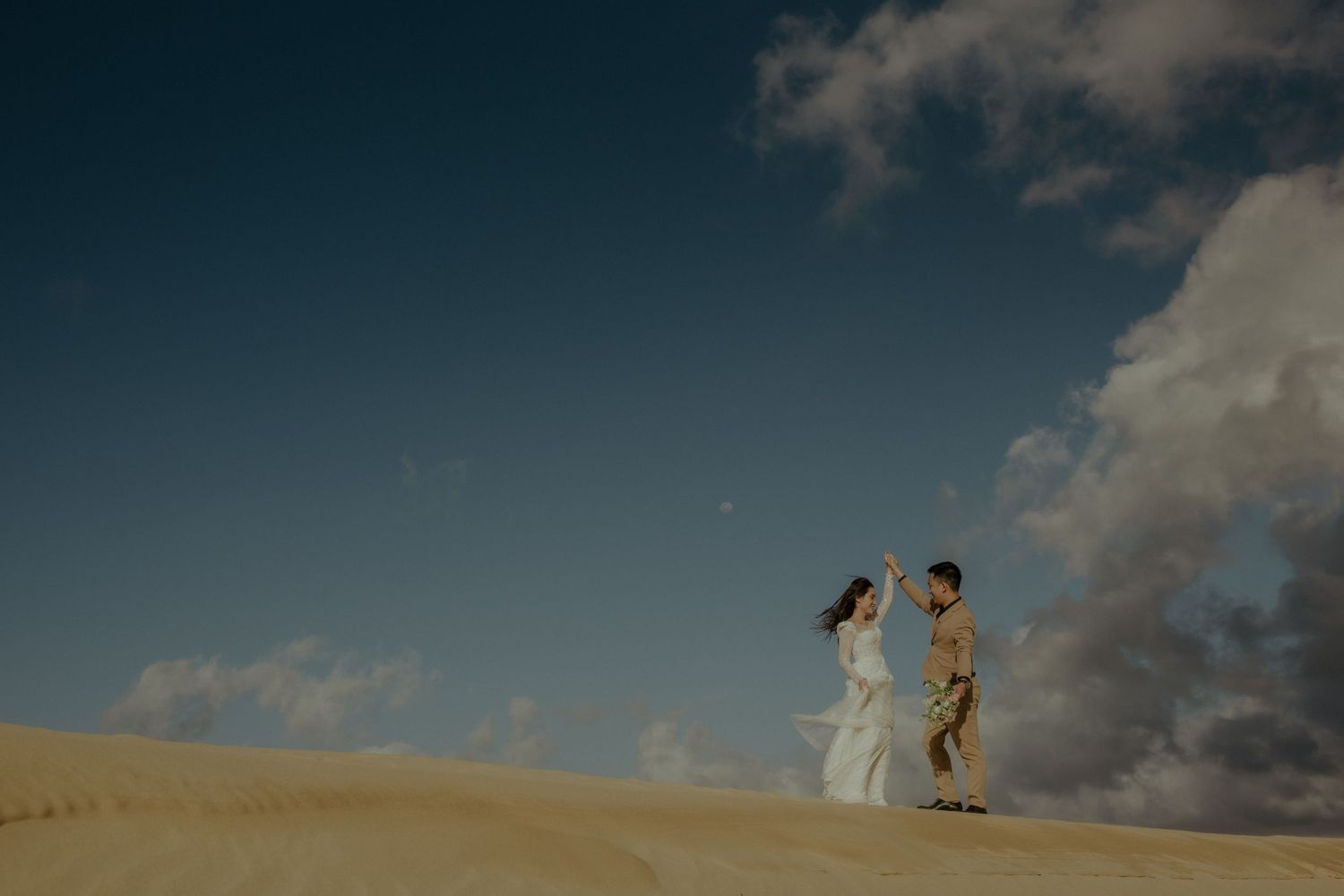 Two figures in elegant attire stand atop a sandy dune against a dramatic blue sky with white clouds at sunset.