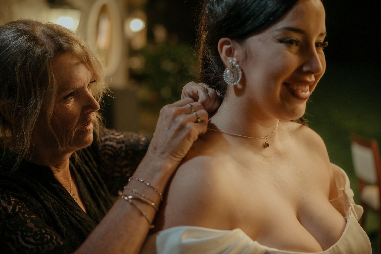 A helper adjusts a white strapless dress in warm lighting during a wedding preparation moment.