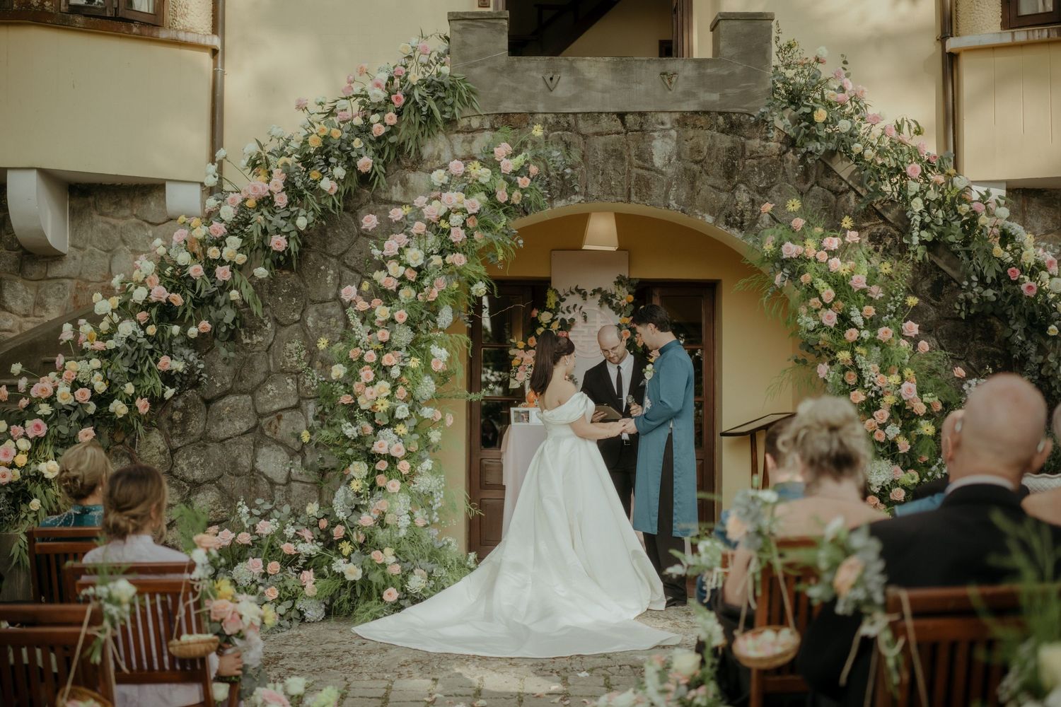 Wedding ceremony under an elegant stone archway decorated with lush white floral garlands at a rustic venue.