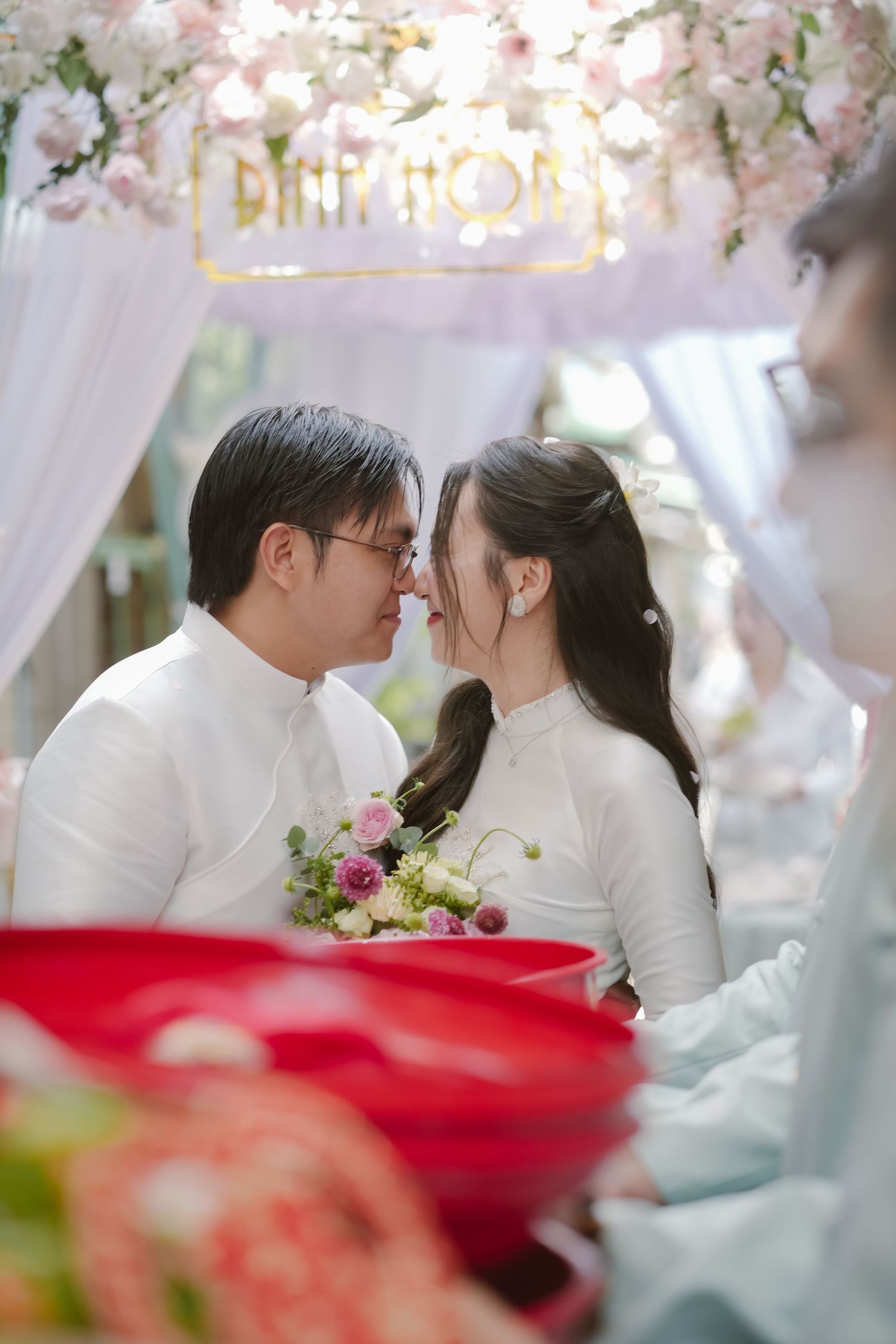 Intimate moment during Vietnamese wedding ceremony under floral arch with pink blossoms.