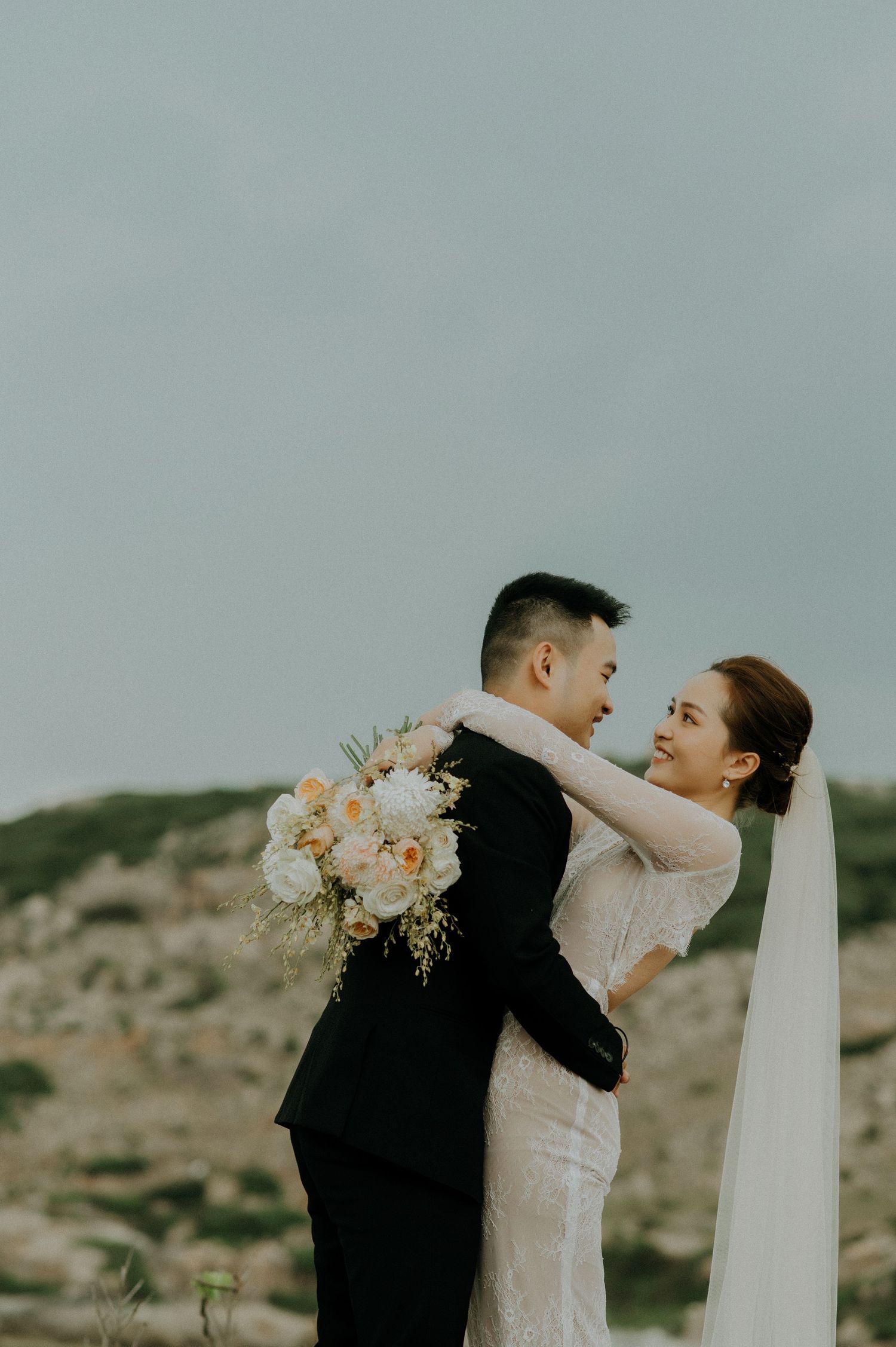 A couple embraces outdoors during their wedding photoshoot with soft peach flowers and flowing veil against a desert landscape.