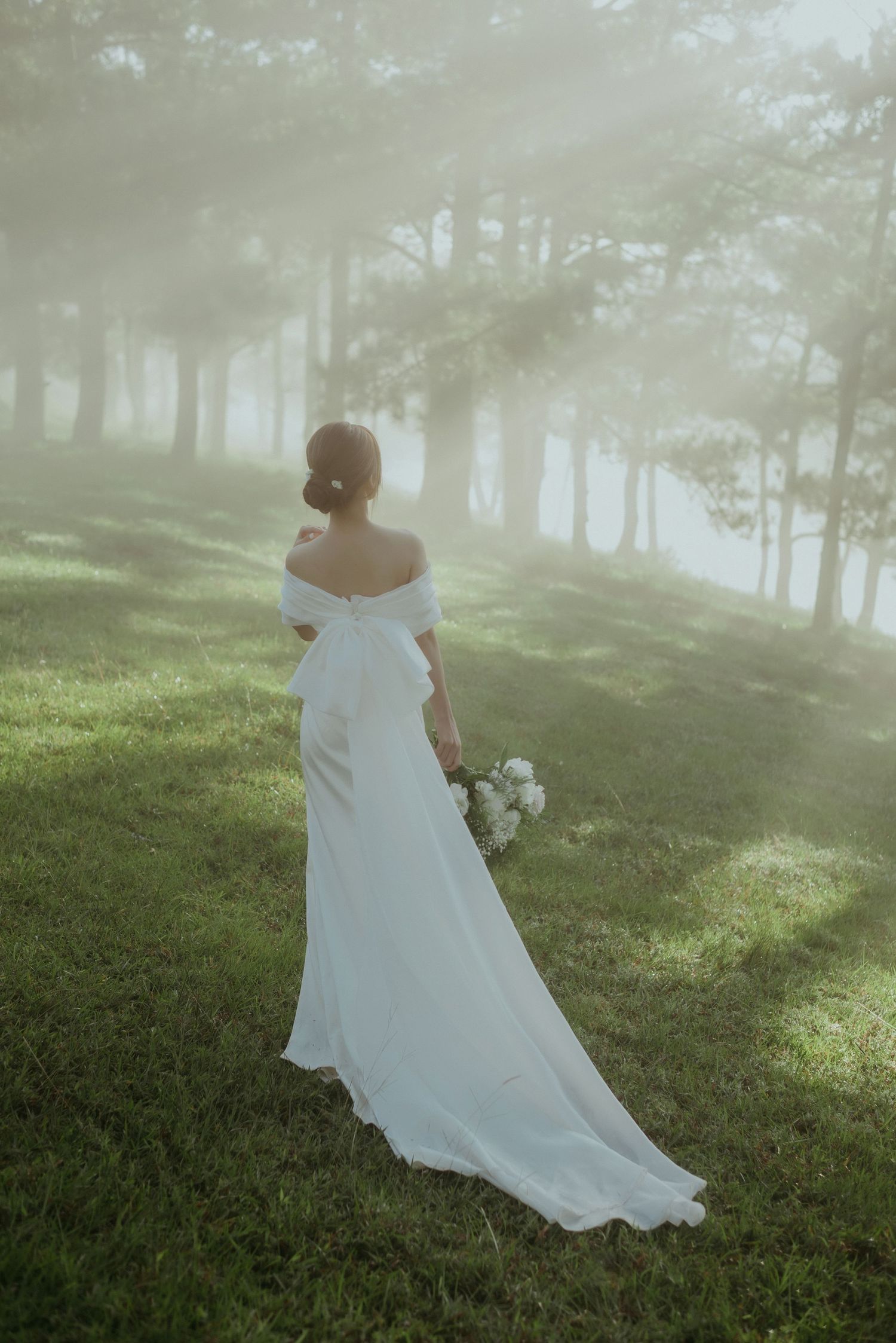 A bride in an elegant white off-shoulder wedding dress stands in a misty forest setting holding a bouquet.