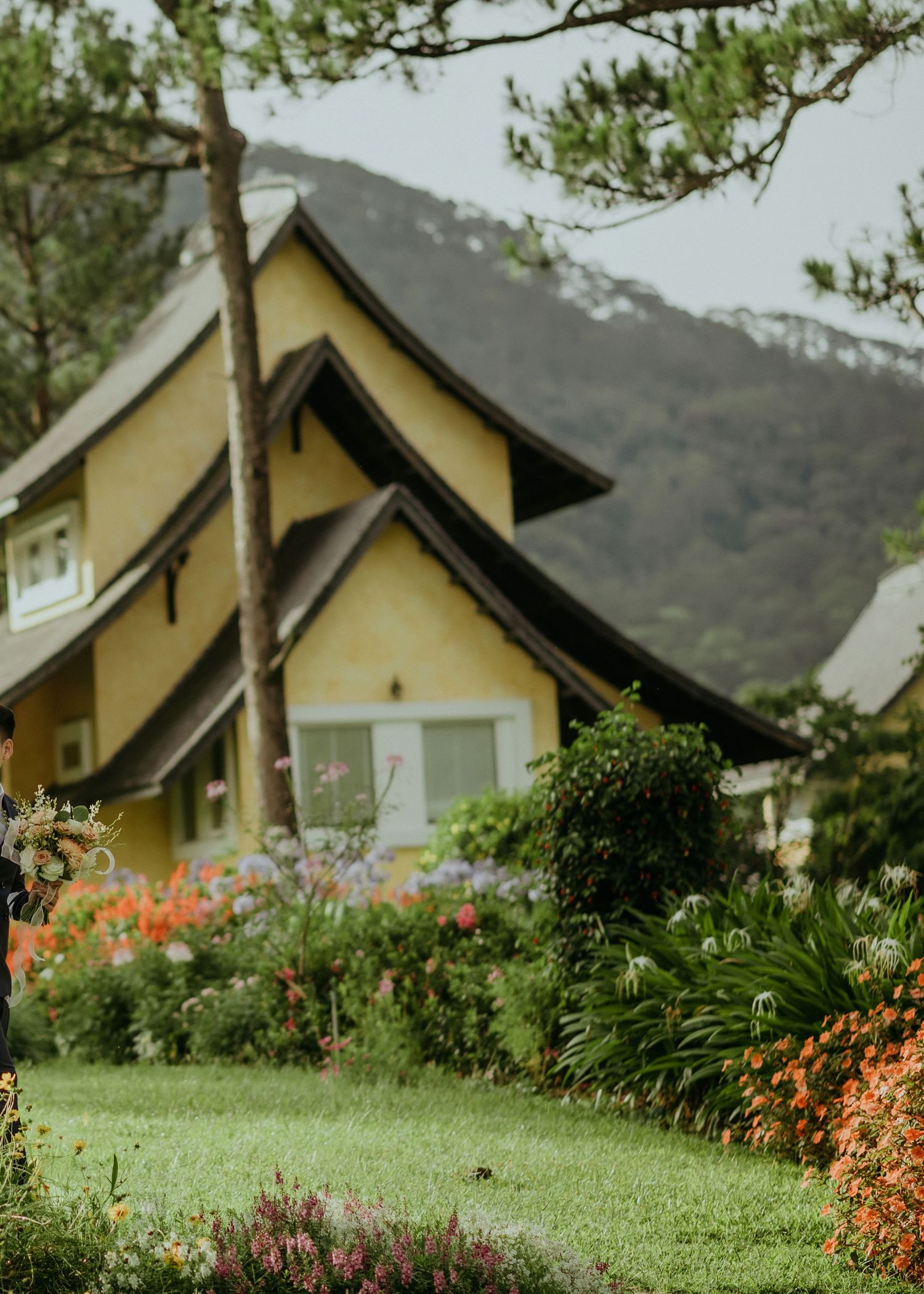 A charming yellow cottage with a steep gabled roof sits nestled in mountains, surrounded by vibrant flower gardens.