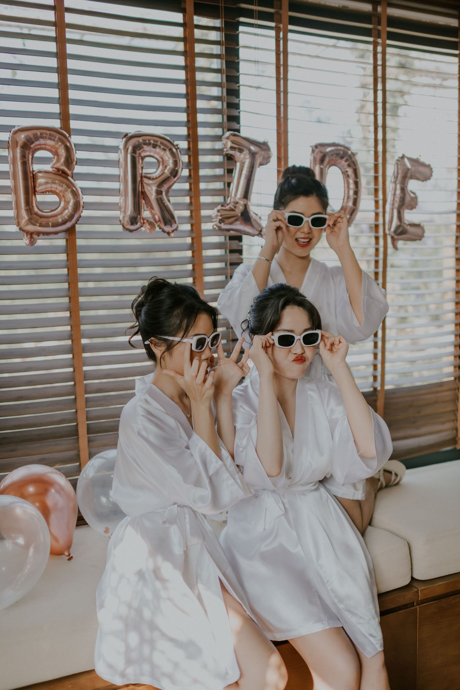 Three people in white robes and sunglasses pose beneath rose gold BRIDE balloon letters.