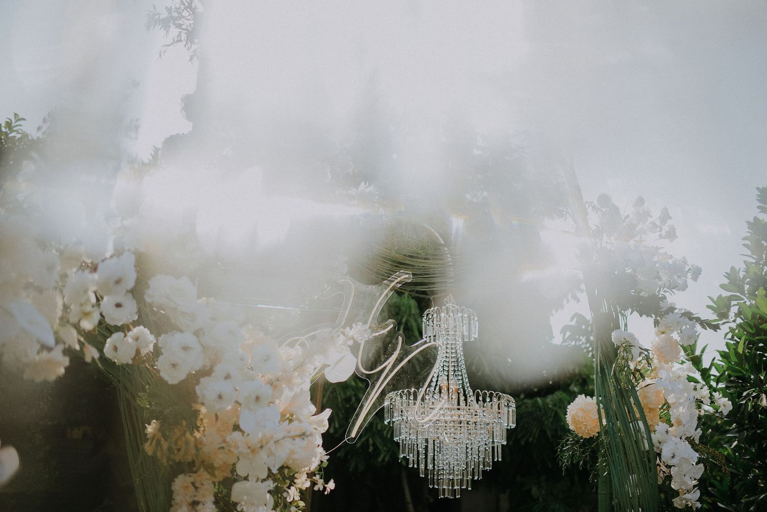 Elegant crystal chandelier hangs beneath white floral arrangements at outdoor wedding ceremony.