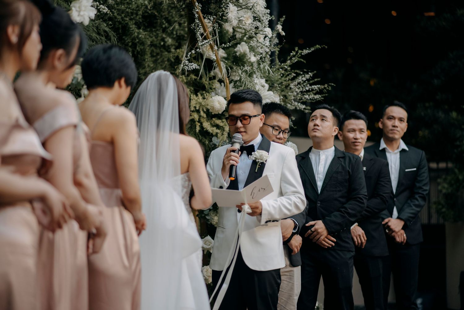 Wedding party wearing pink bridesmaids dresses and black suits gather during outdoor ceremony with floral decor.