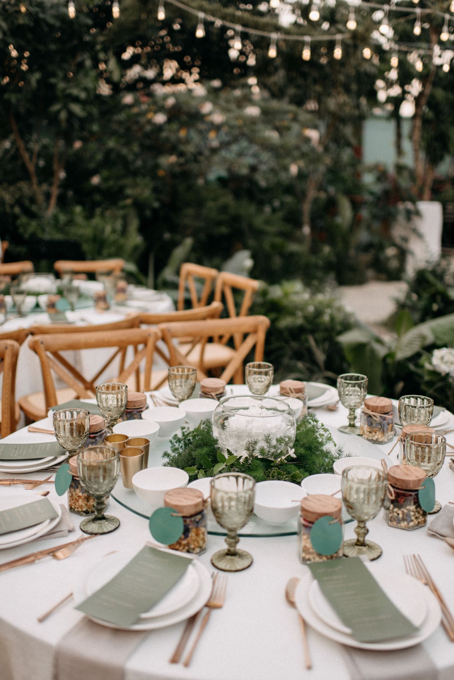 Elegant wedding table setting with crystal glasses, white linens, and greenery centerpiece under string lights.