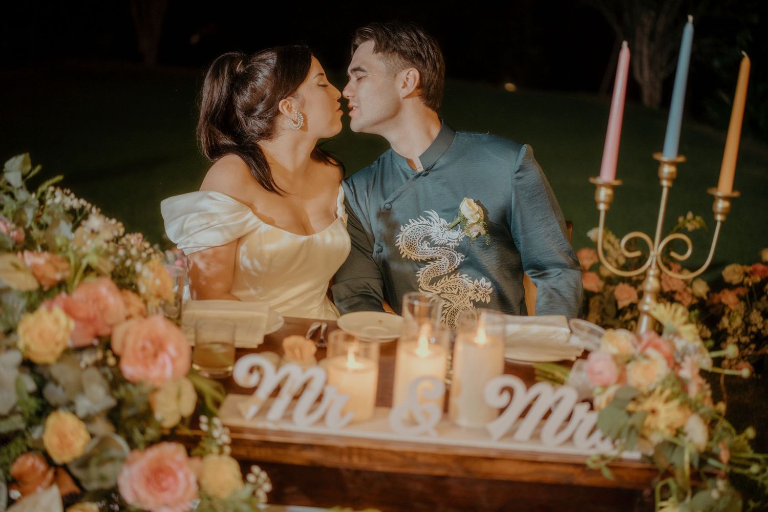 Elegant wedding table setup with gold candelabra, soft pink floral arrangements, and Mr & Mrs sign in romantic evening lighting.