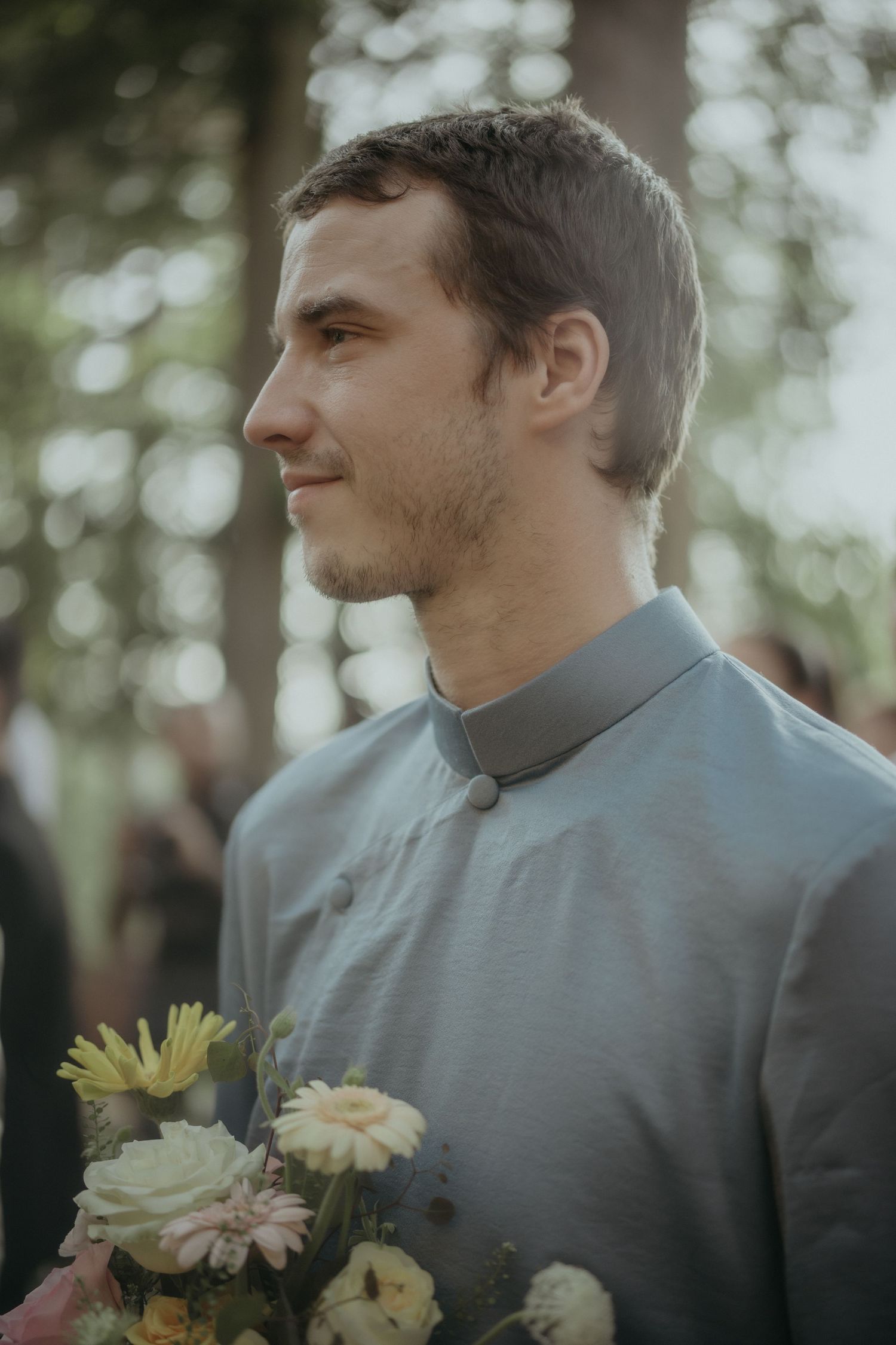 A person in a gray mandarin collar shirt holds a bouquet of yellow and white flowers while looking to the side outdoors.