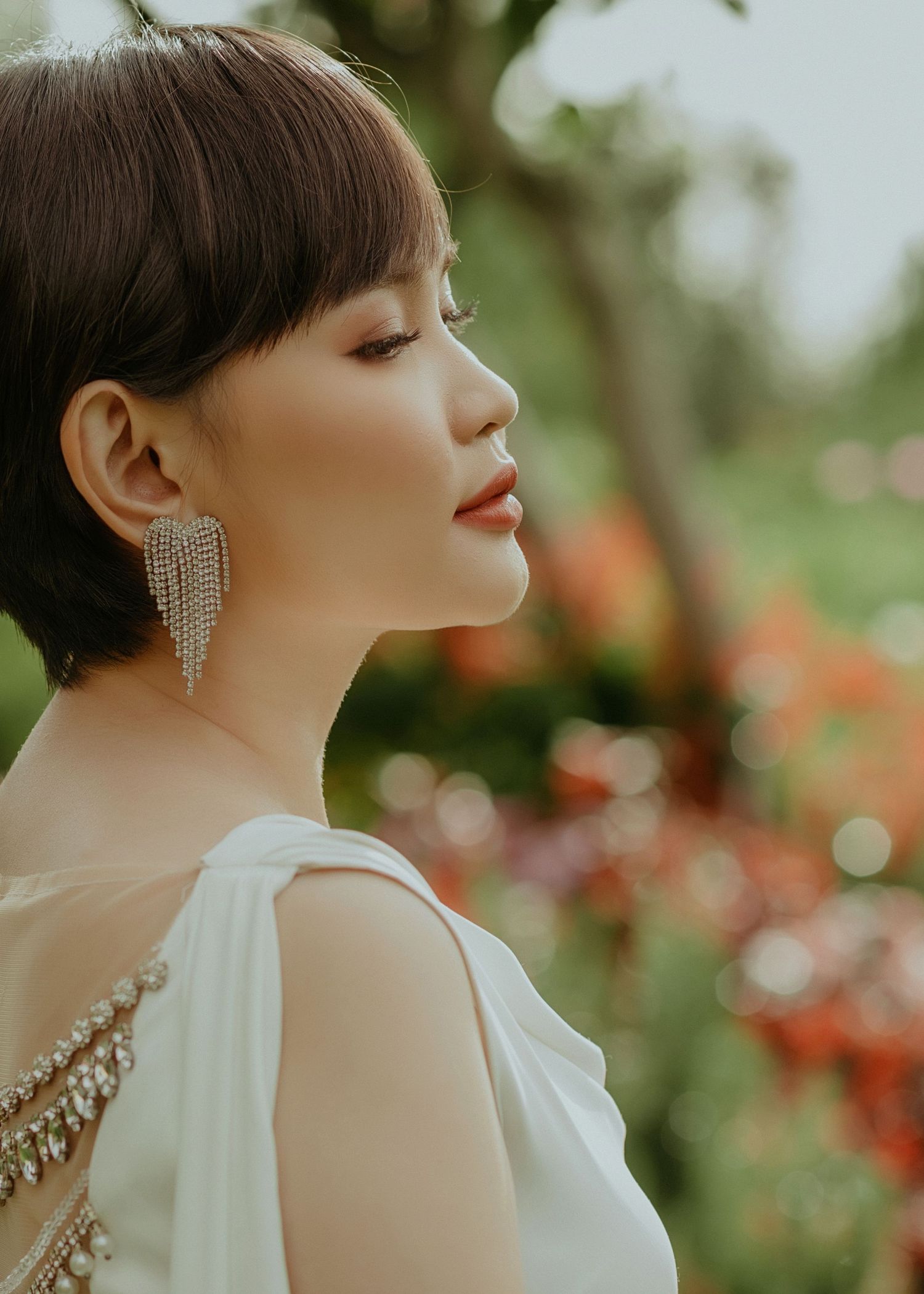 Side profile portrait in white dress with sparkly earrings against blurred garden backdrop with red flowers.