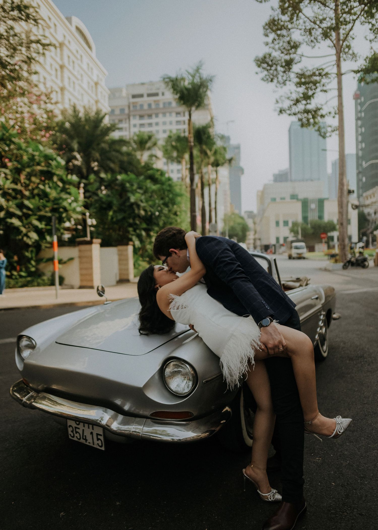A romantic scene featuring a couple sharing a kiss while leaning back on a classic silver automobile on a city street.