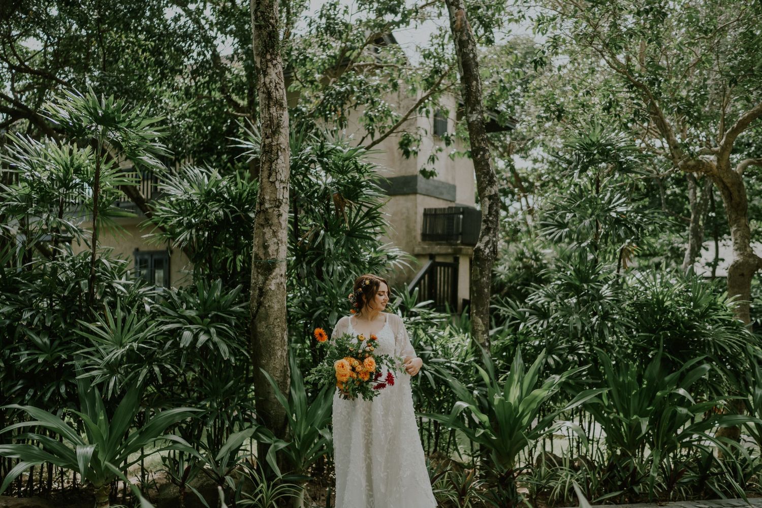 A bride in a lace wedding dress carries a vibrant orange and red bouquet while walking through a lush tropical garden setting.