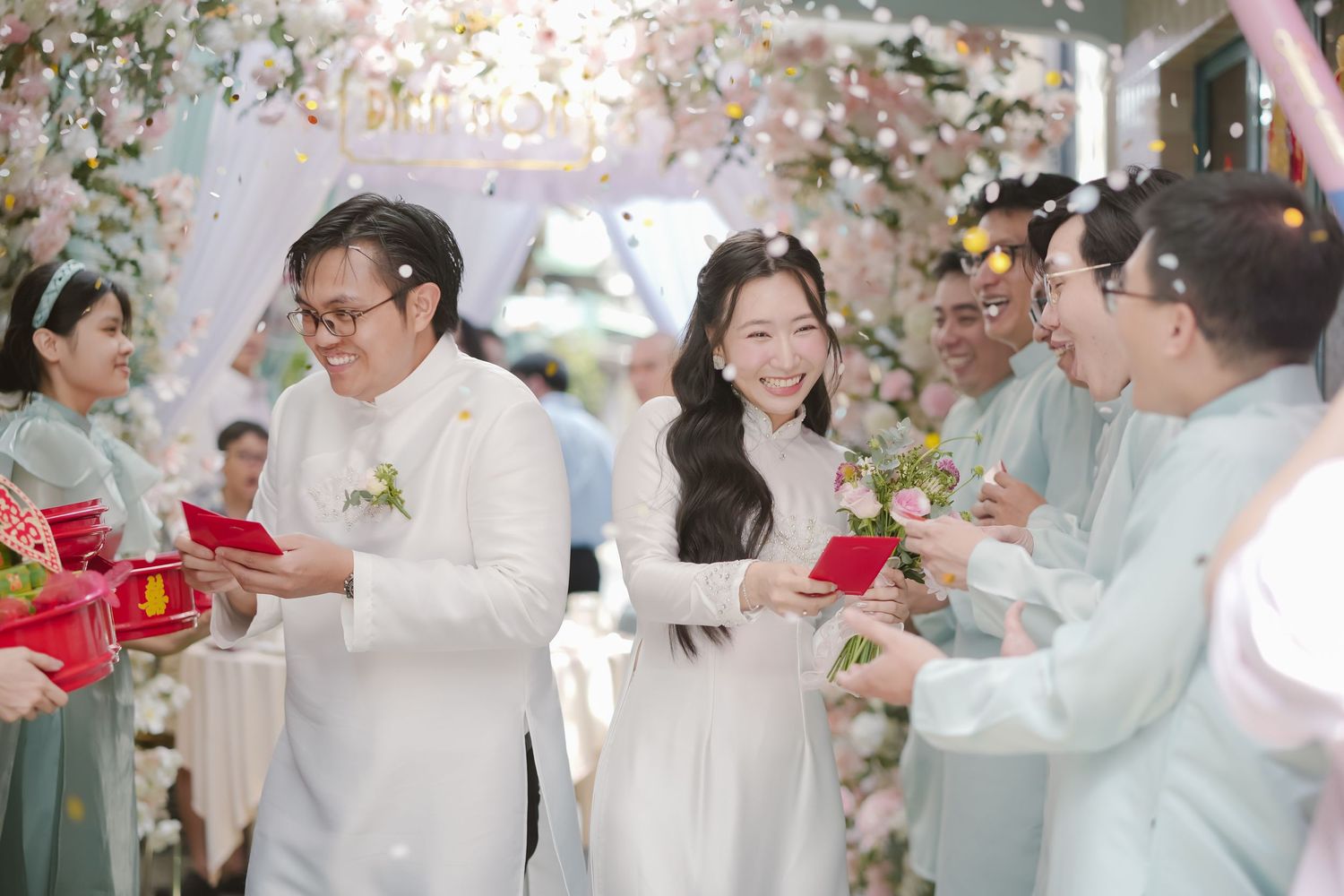Wedding celebration with guests in white and mint green attire sharing gifts under floral decorations.