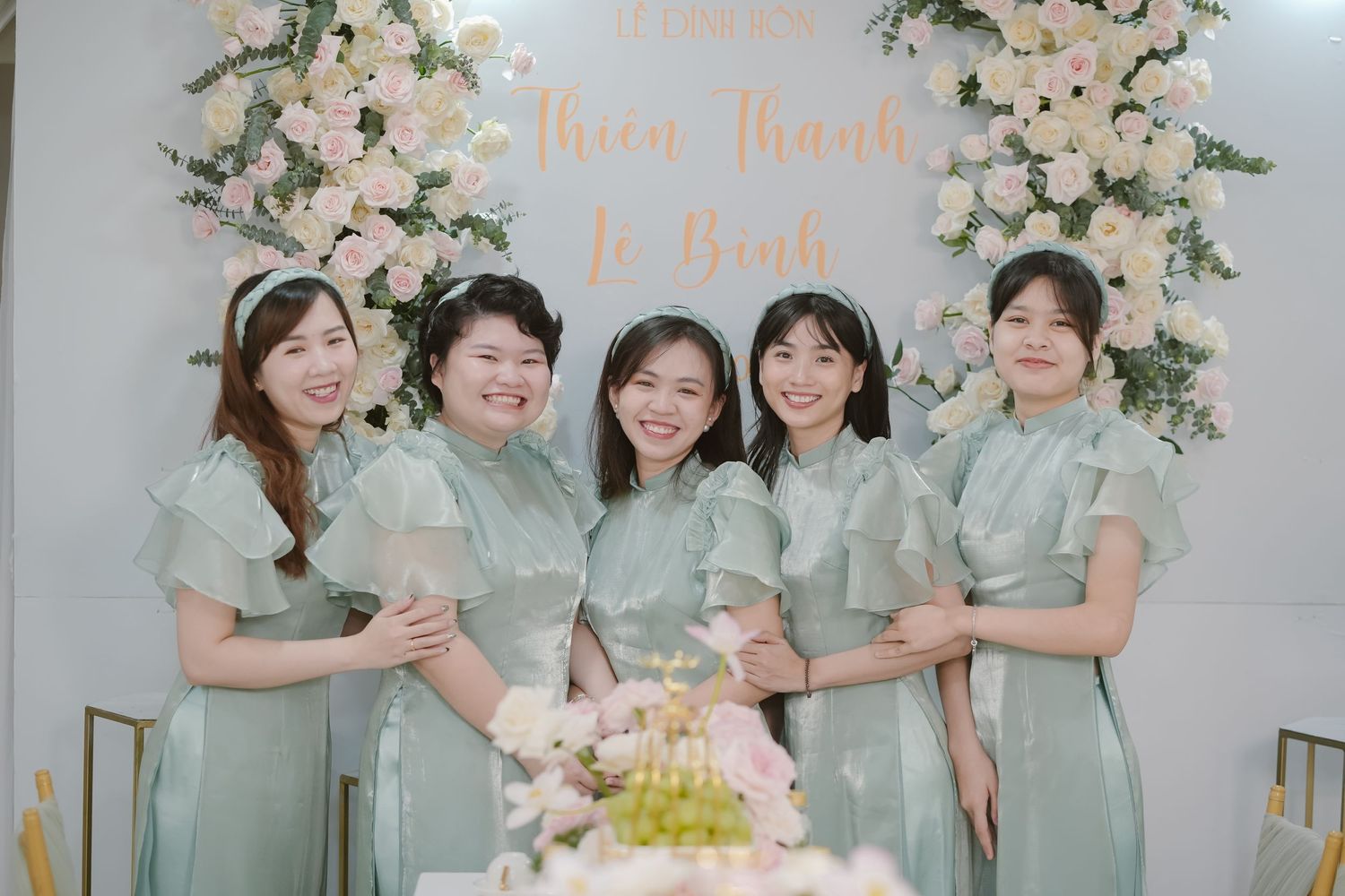A group wearing mint green dresses poses in front of a floral arch at a wedding celebration.