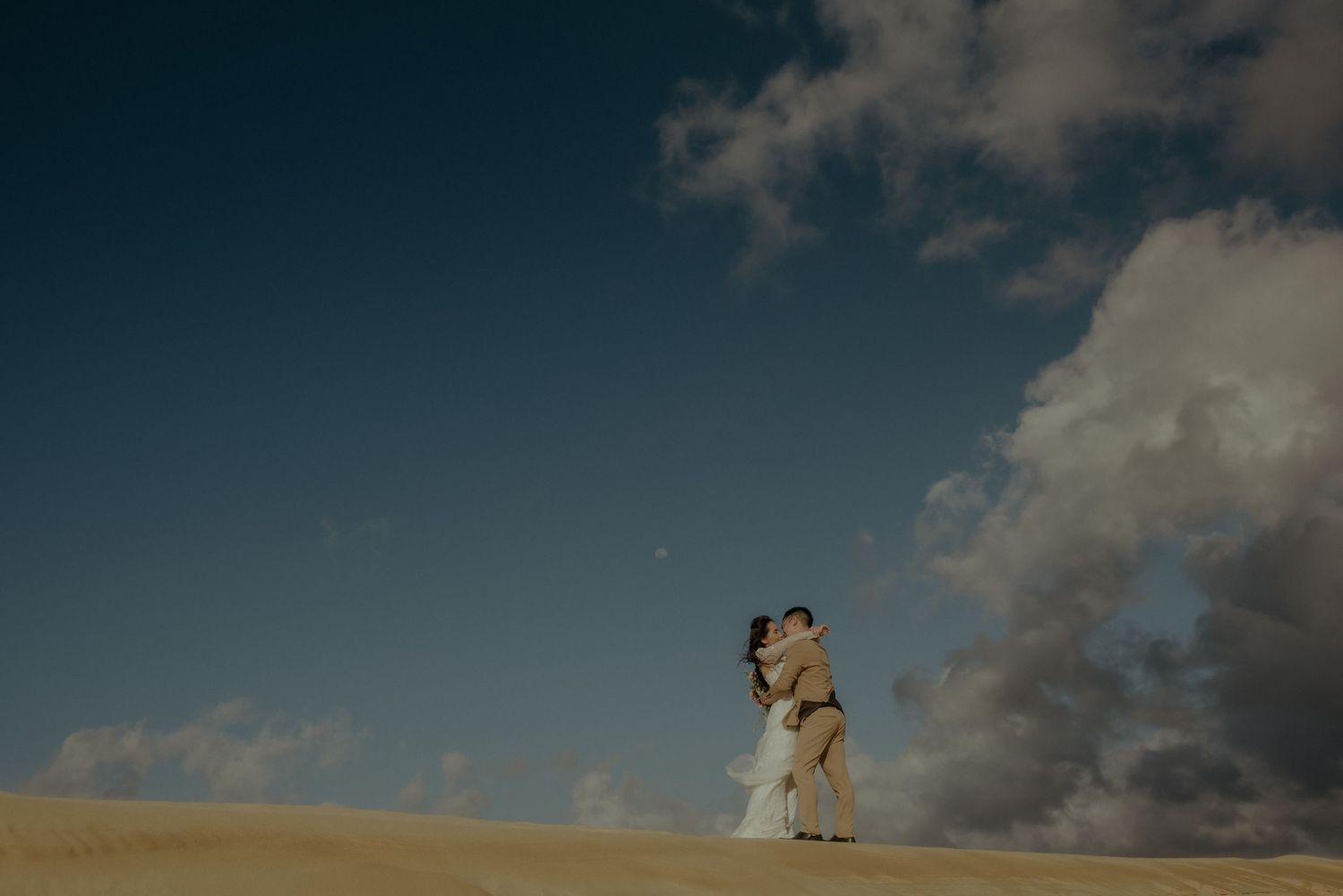 Two figures stand silhouetted against a dramatic sky with clouds on a sand dune during golden hour.