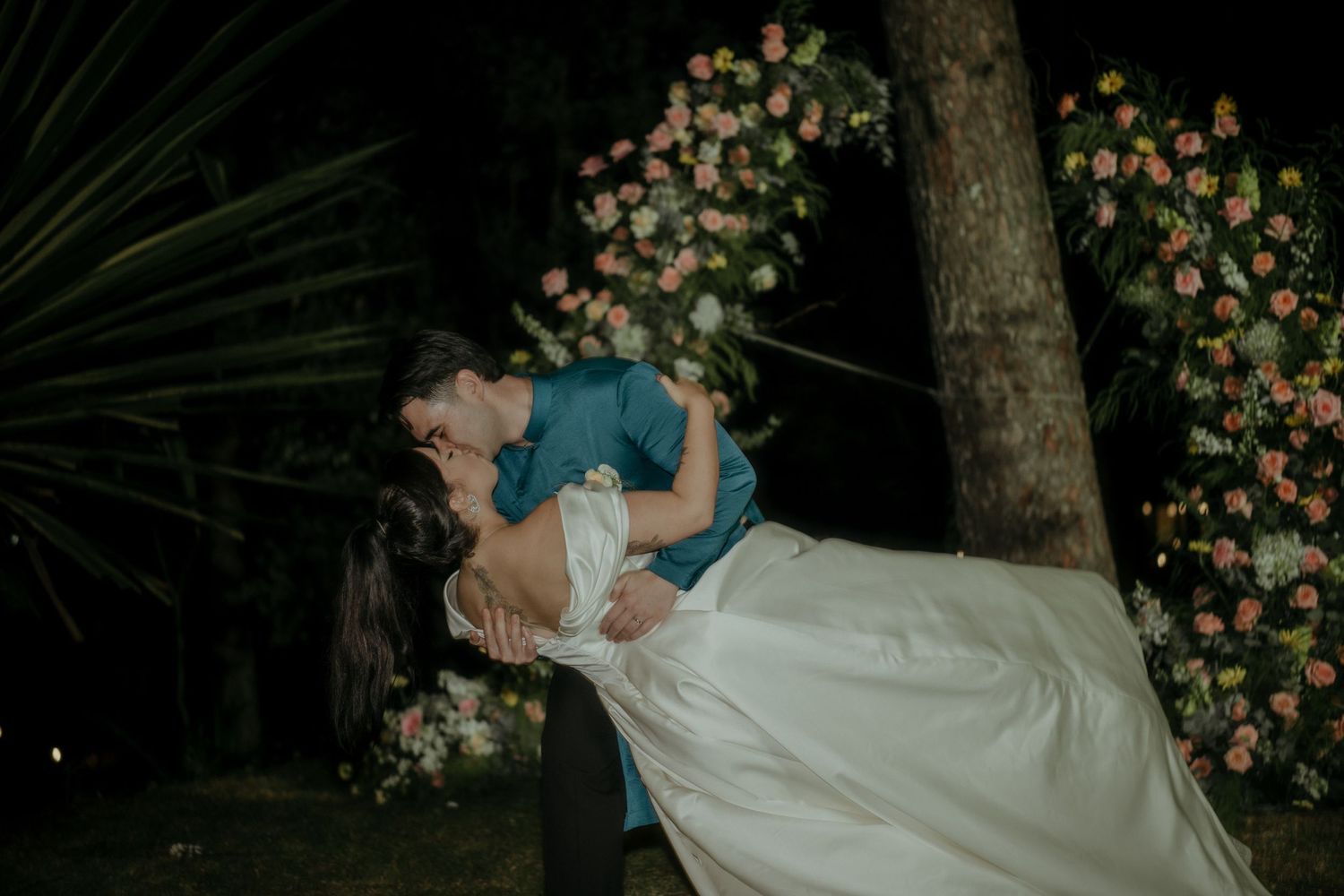 A romantic dip kiss at night in a garden with blooming pink flowers and palm trees in the background.