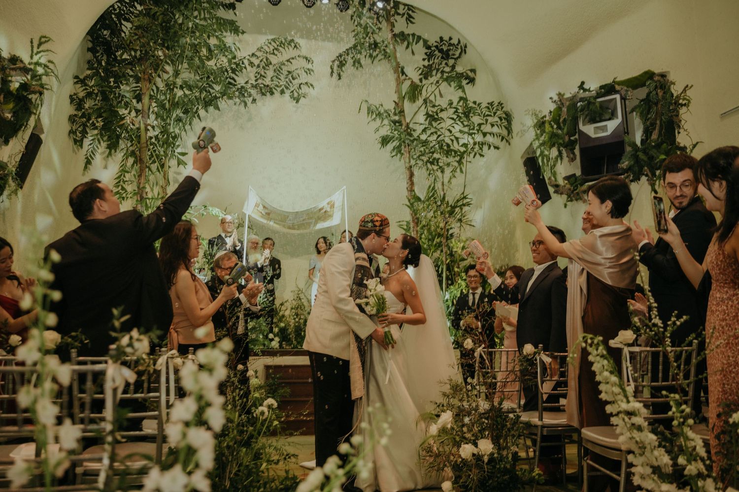 Wedding guests throw confetti and cheer during an indoor garden-themed ceremony.